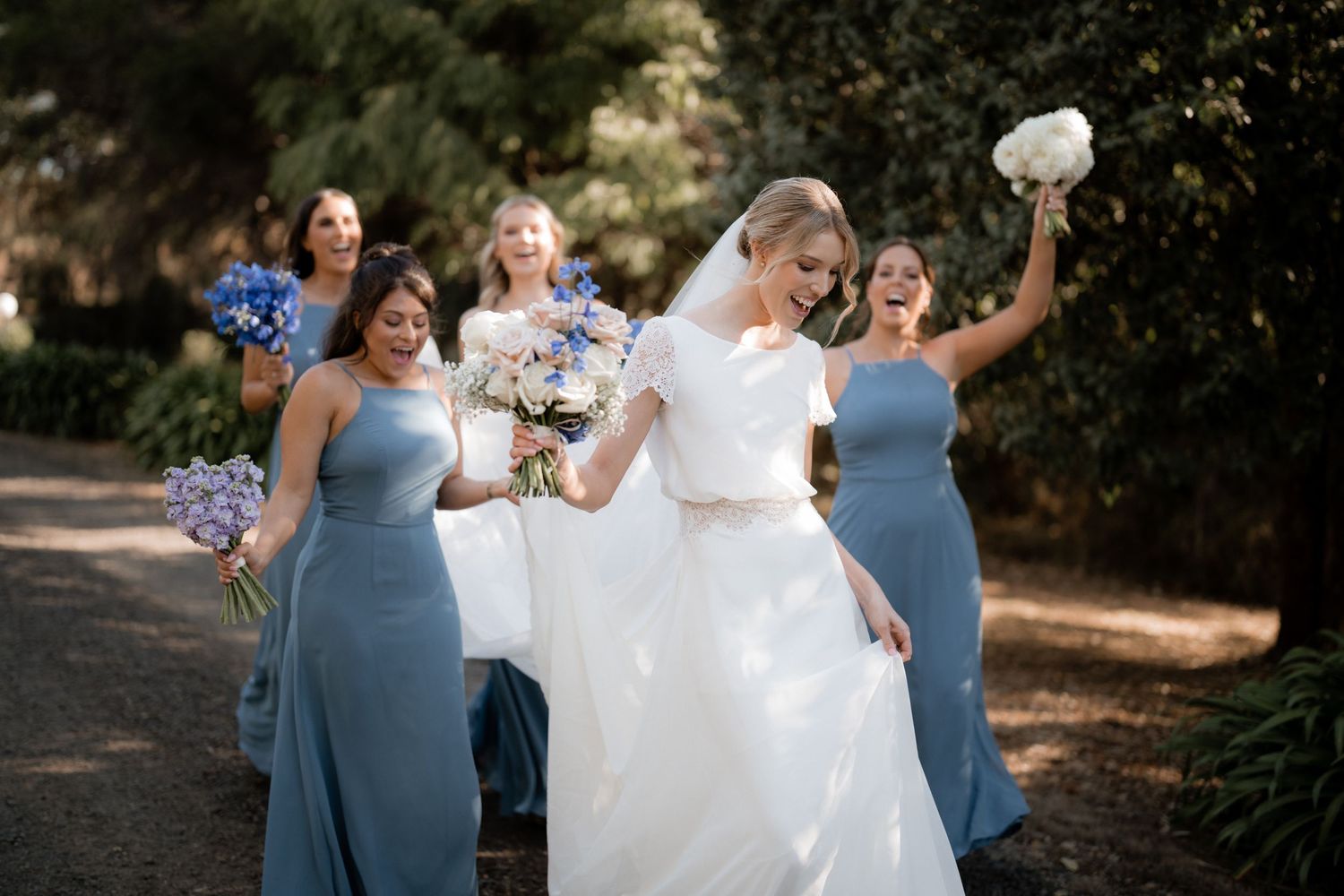 A bridal party celebrates on a garden path with bridesmaids in blue dresses and bouquets.