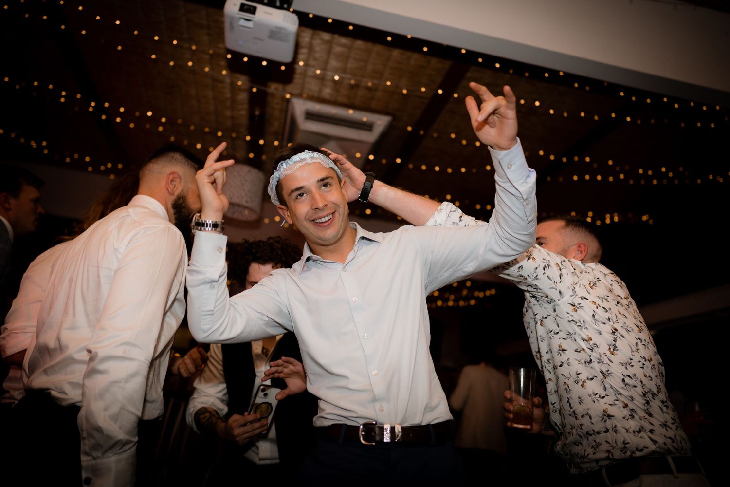People dancing and celebrating at a wedding reception under string lights with energetic movement captured in dim lighting.