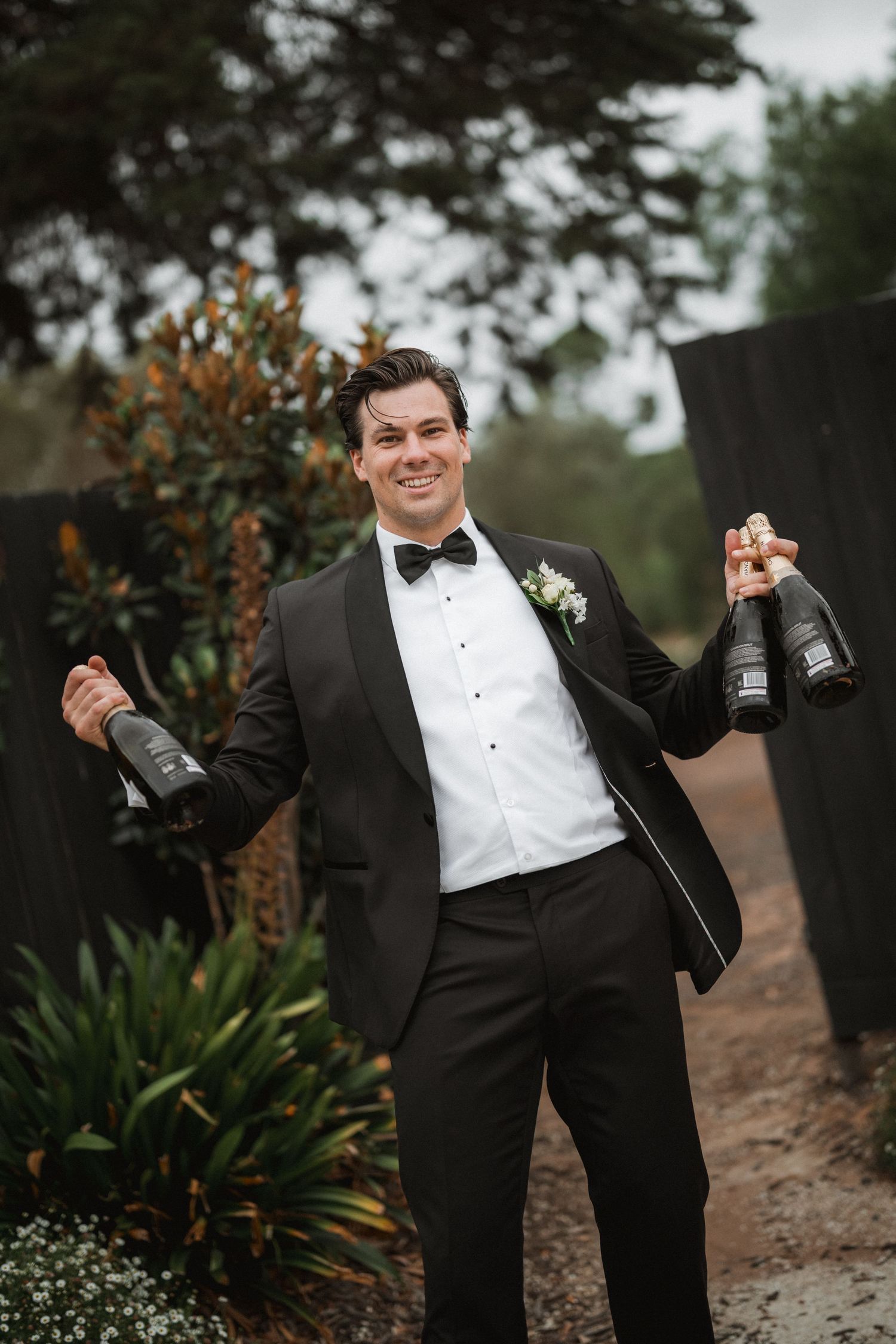 Groomsman in tuxedo celebrating with champagne bottles outdoors.