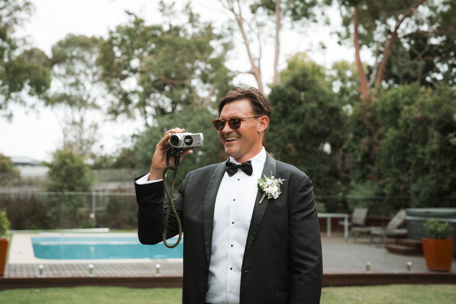 Someone in a black tuxedo and bow tie holds binoculars while standing outdoors near a pool.