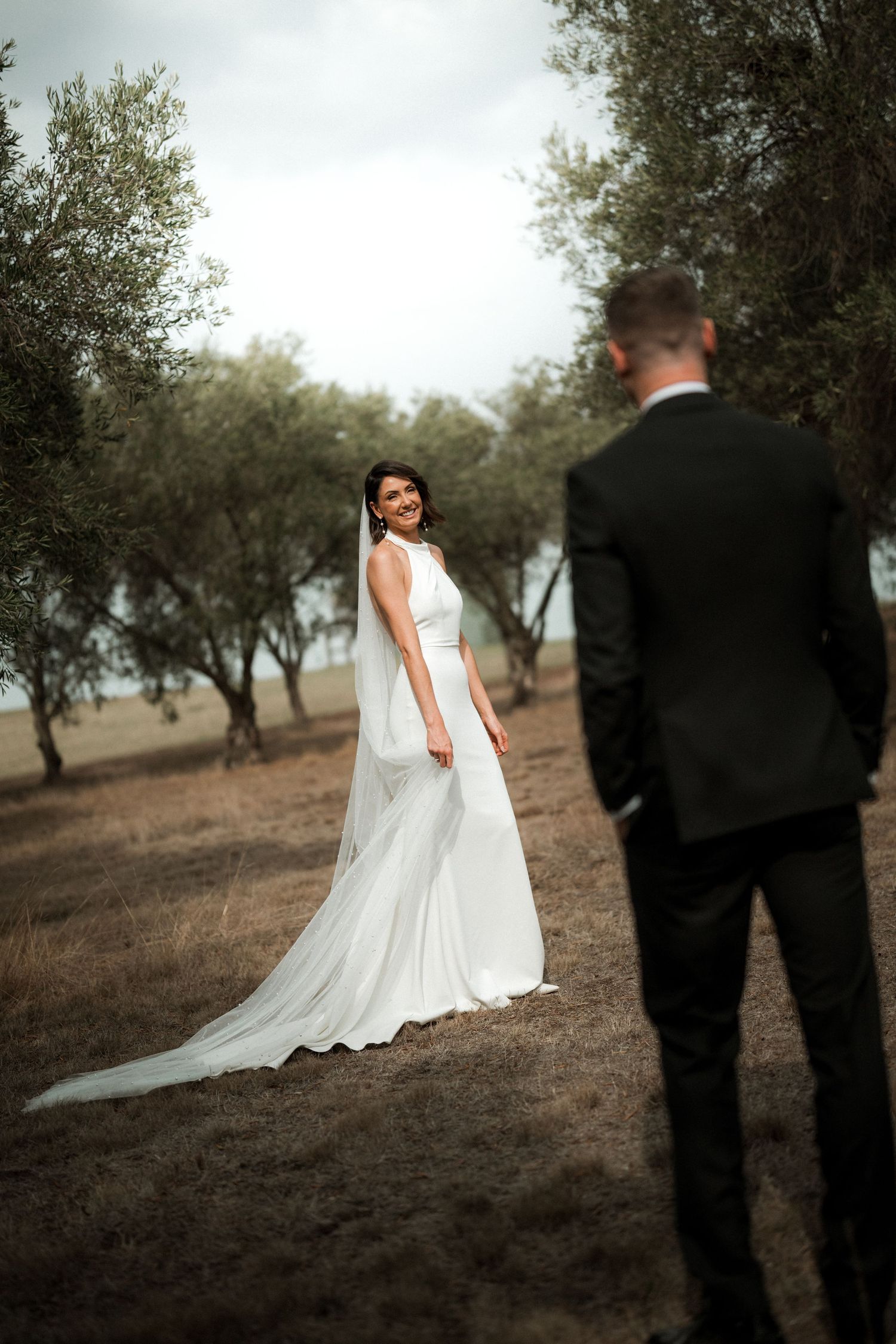 A bride in a flowing white gown poses for a first look photo with her groom in a rustic outdoor setting.