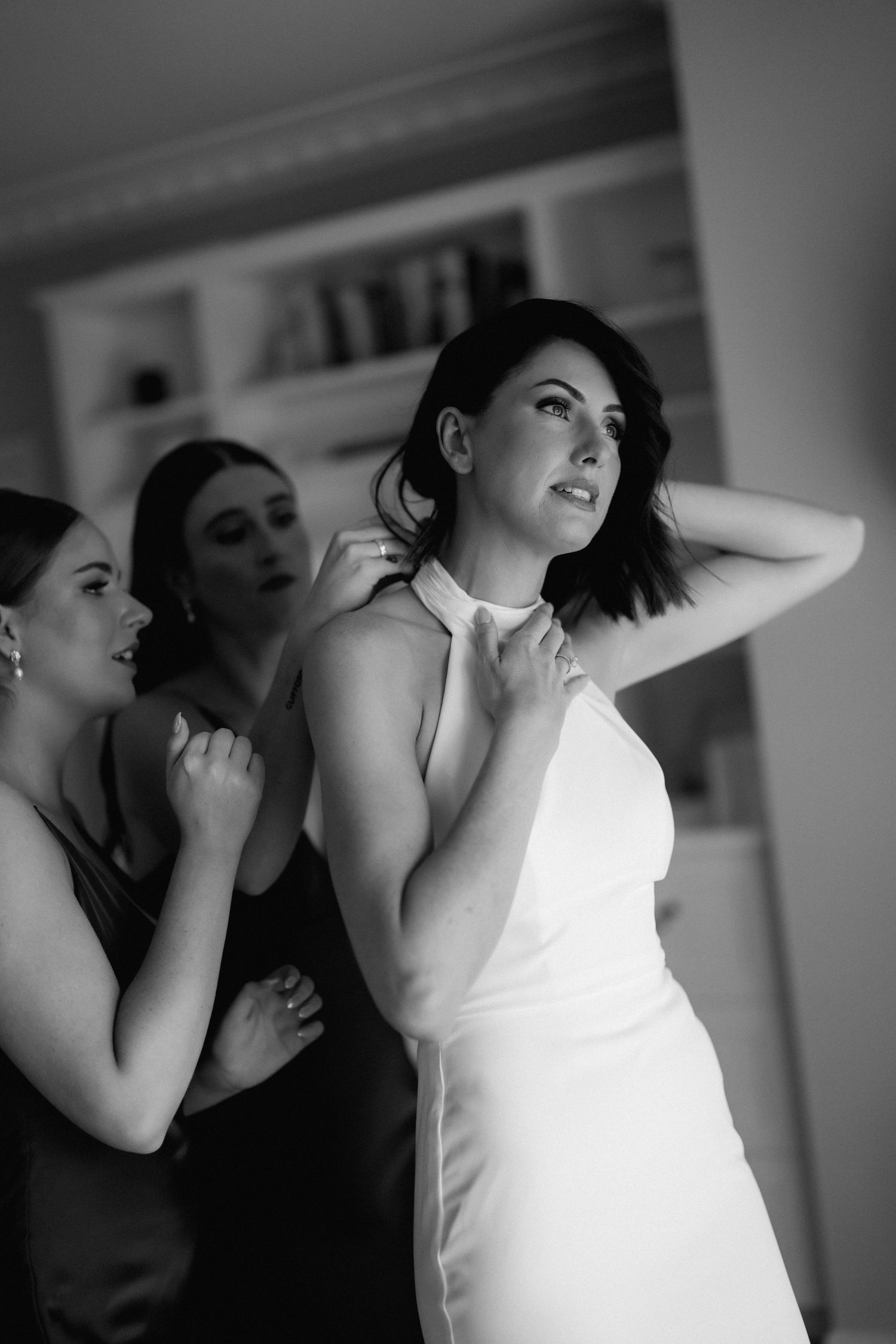 Bridesmaids help a bride in an elegant white dress get ready in a candid black and white wedding preparation moment.