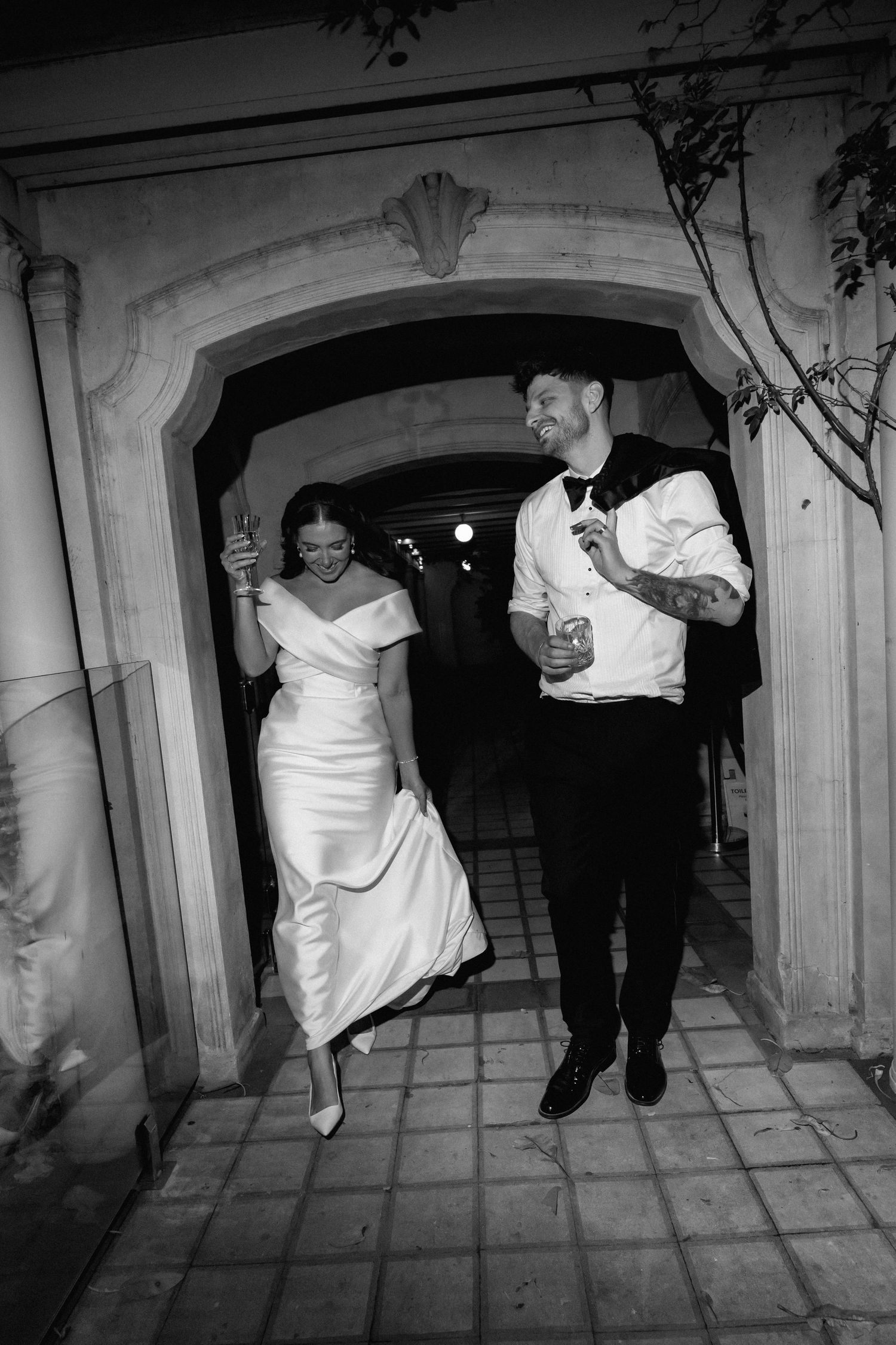 A bride and groom exit through an arched doorway at night, both holding drinks and smiling while walking together.
