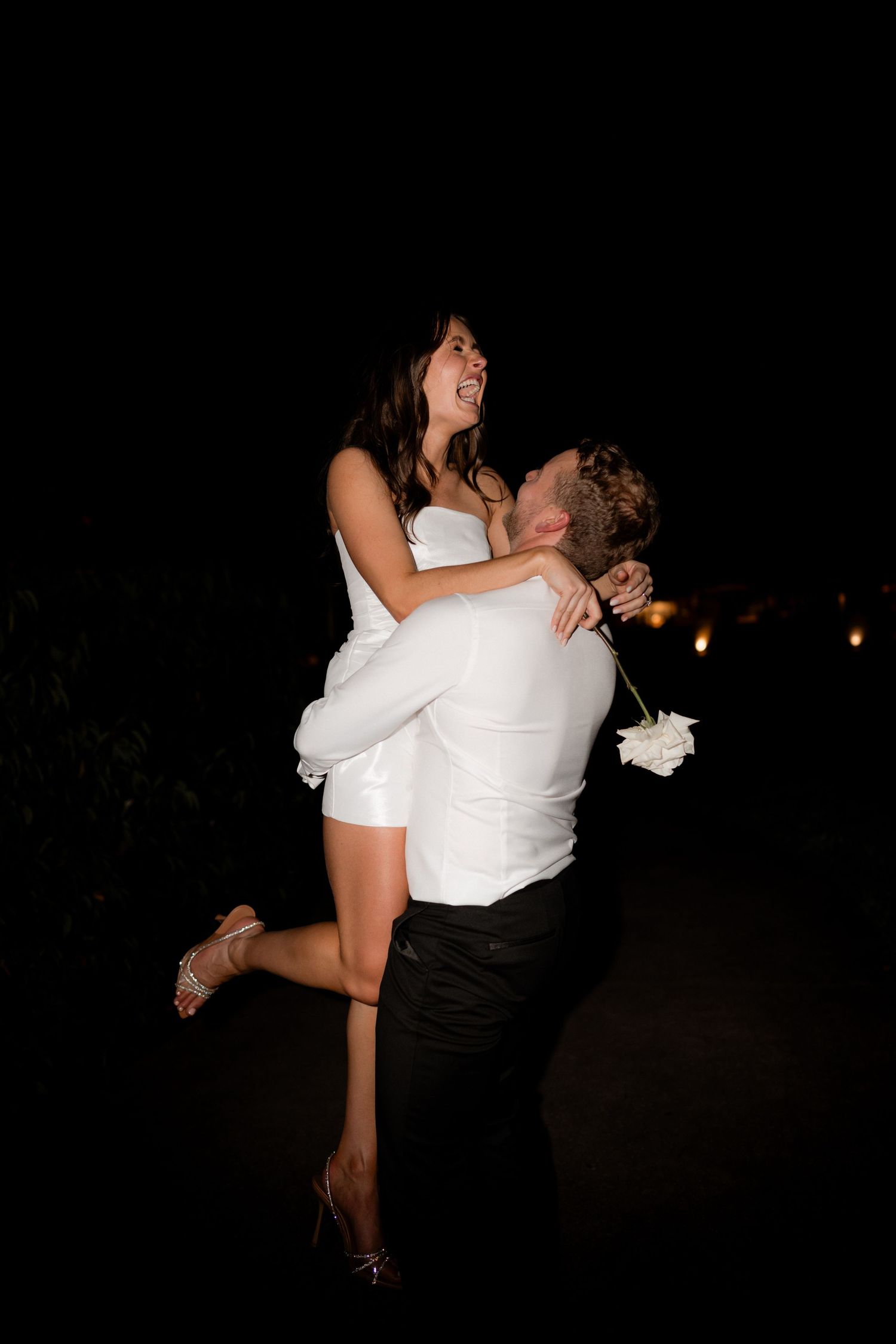 A couple shares a romantic dance pose in formal white attire at a nighttime celebration.