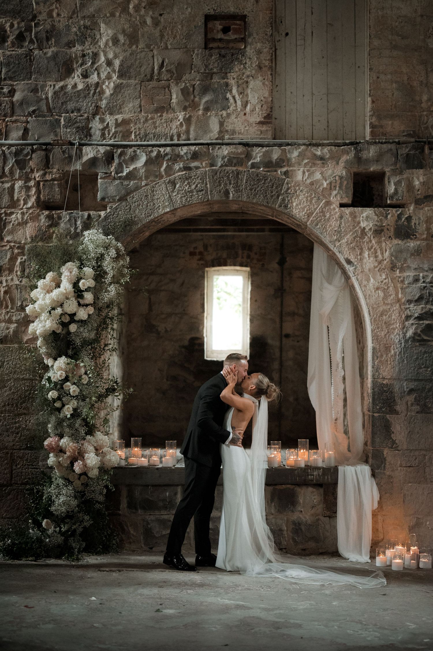 Romantic wedding scene with white floral arch and glowing candles in a stone archway.