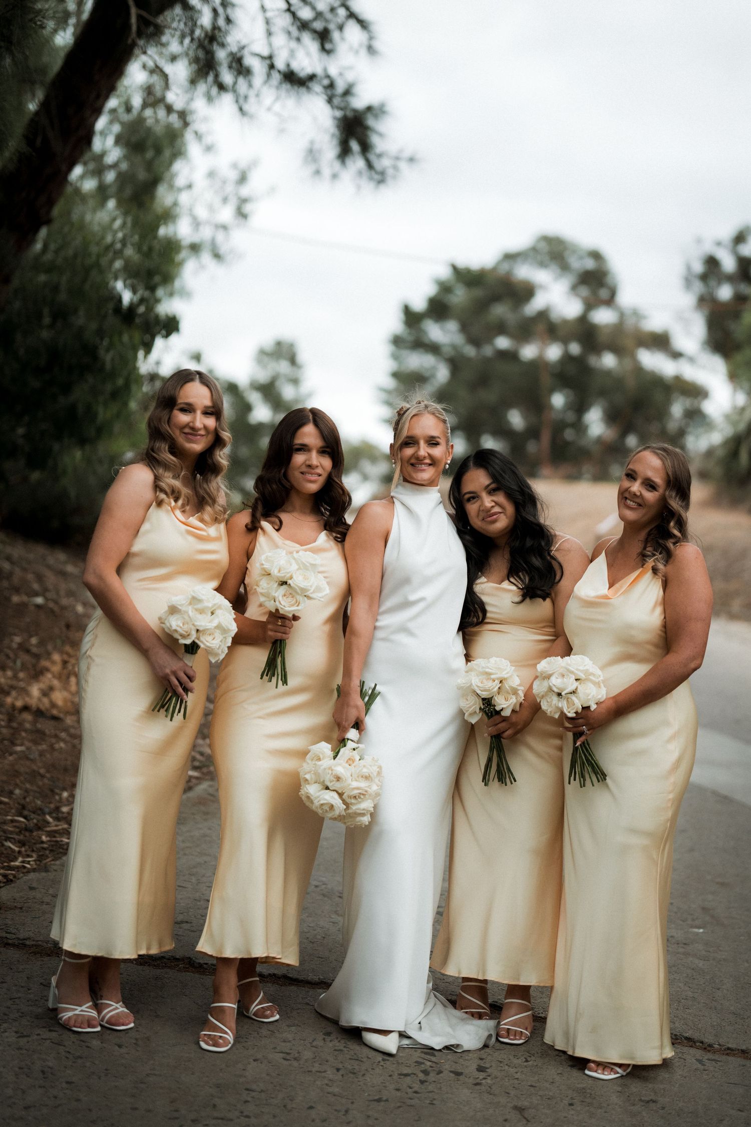 Wedding party photo featuring bridesmaids in cream-colored dresses holding white bouquets on a tree-lined path.