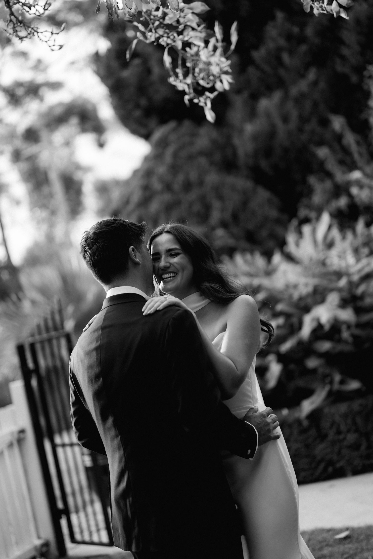Black and white romantic moment captured between a couple in formal attire under garden trees.