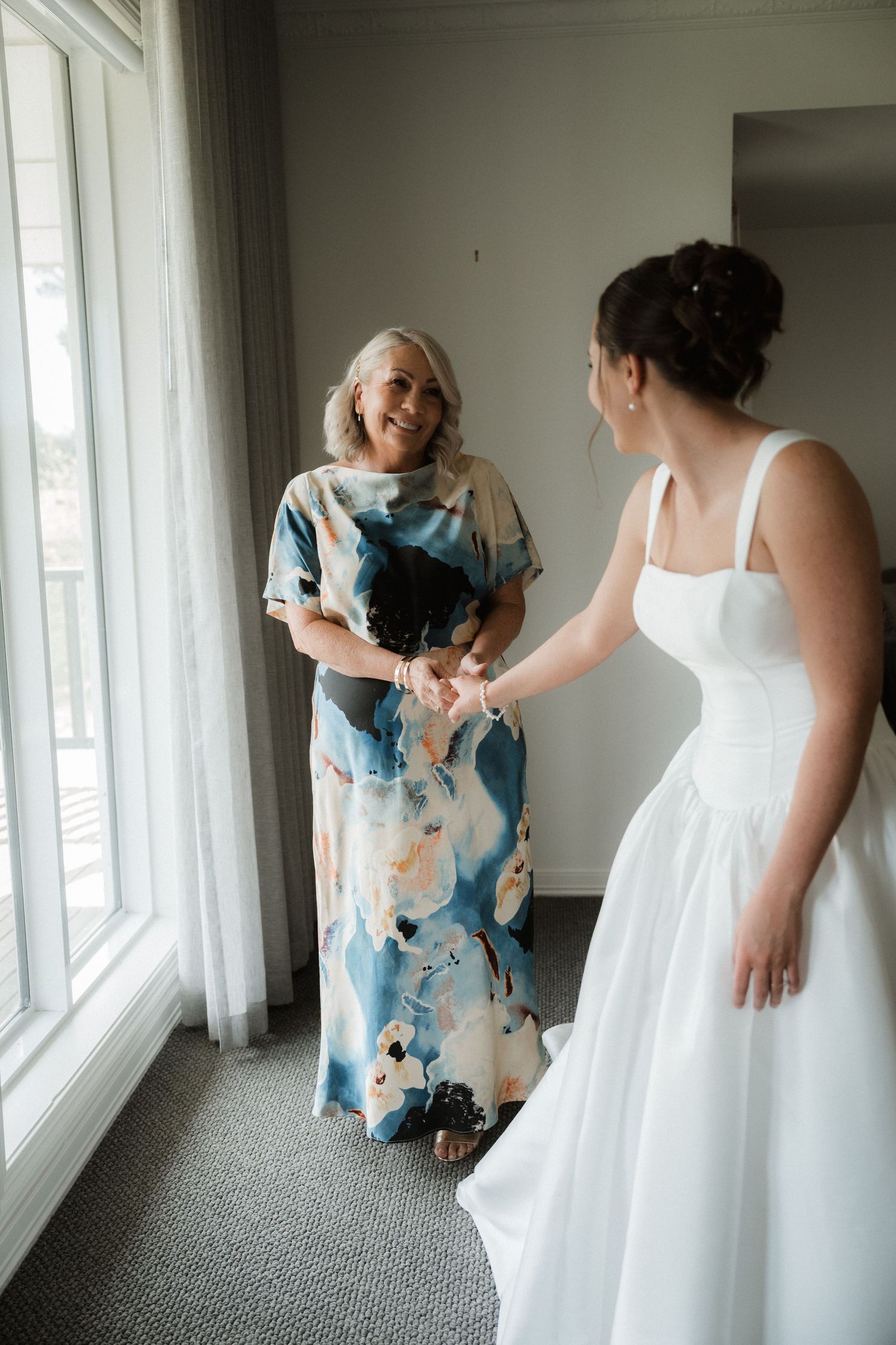 Wedding preparation scene showing a bride in white dress getting ready in a bright room with floor-length windows.
