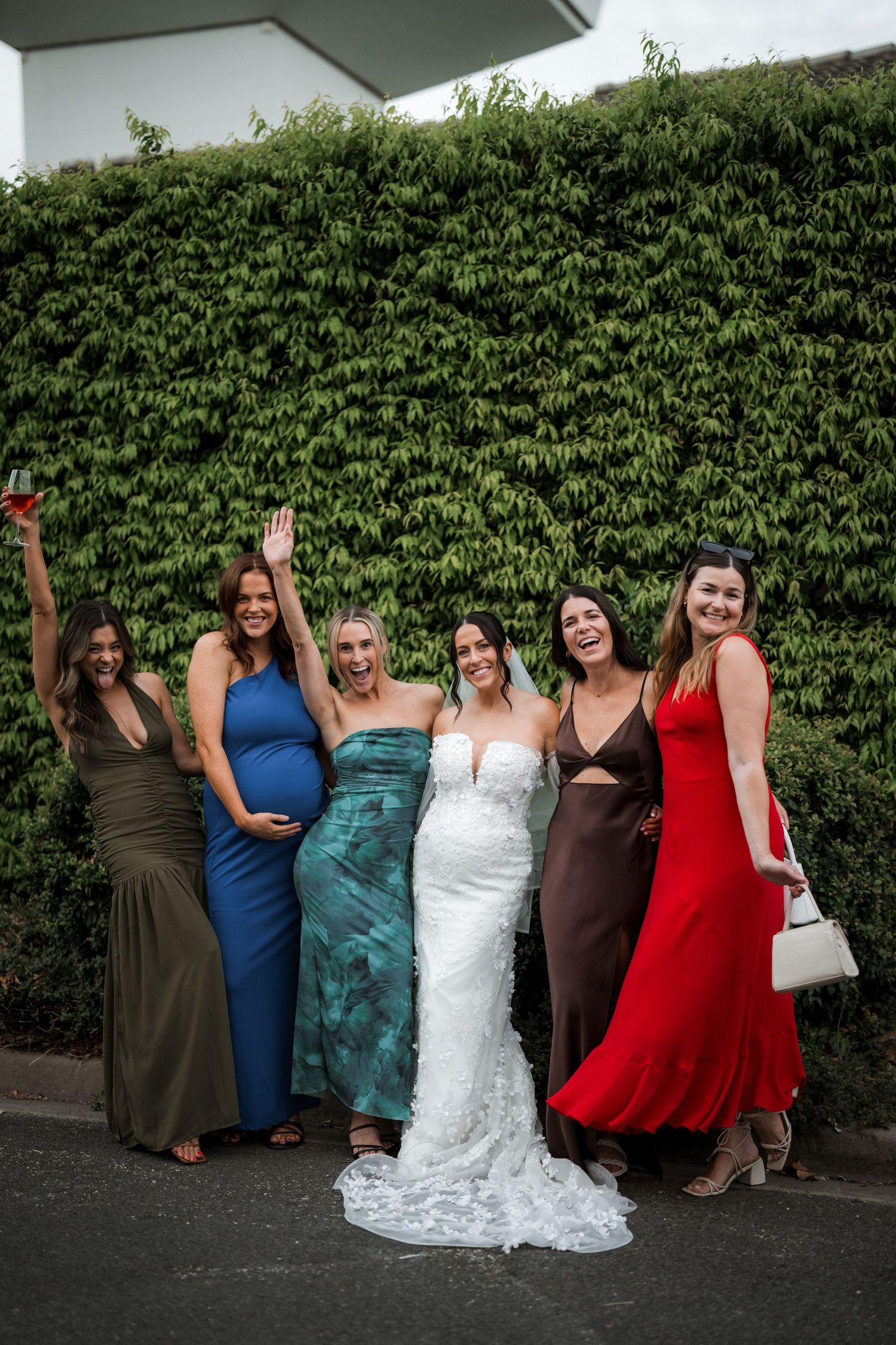Group of bridesmaids in colorful dresses posing with the bride in a lace wedding gown against a hedge wall.