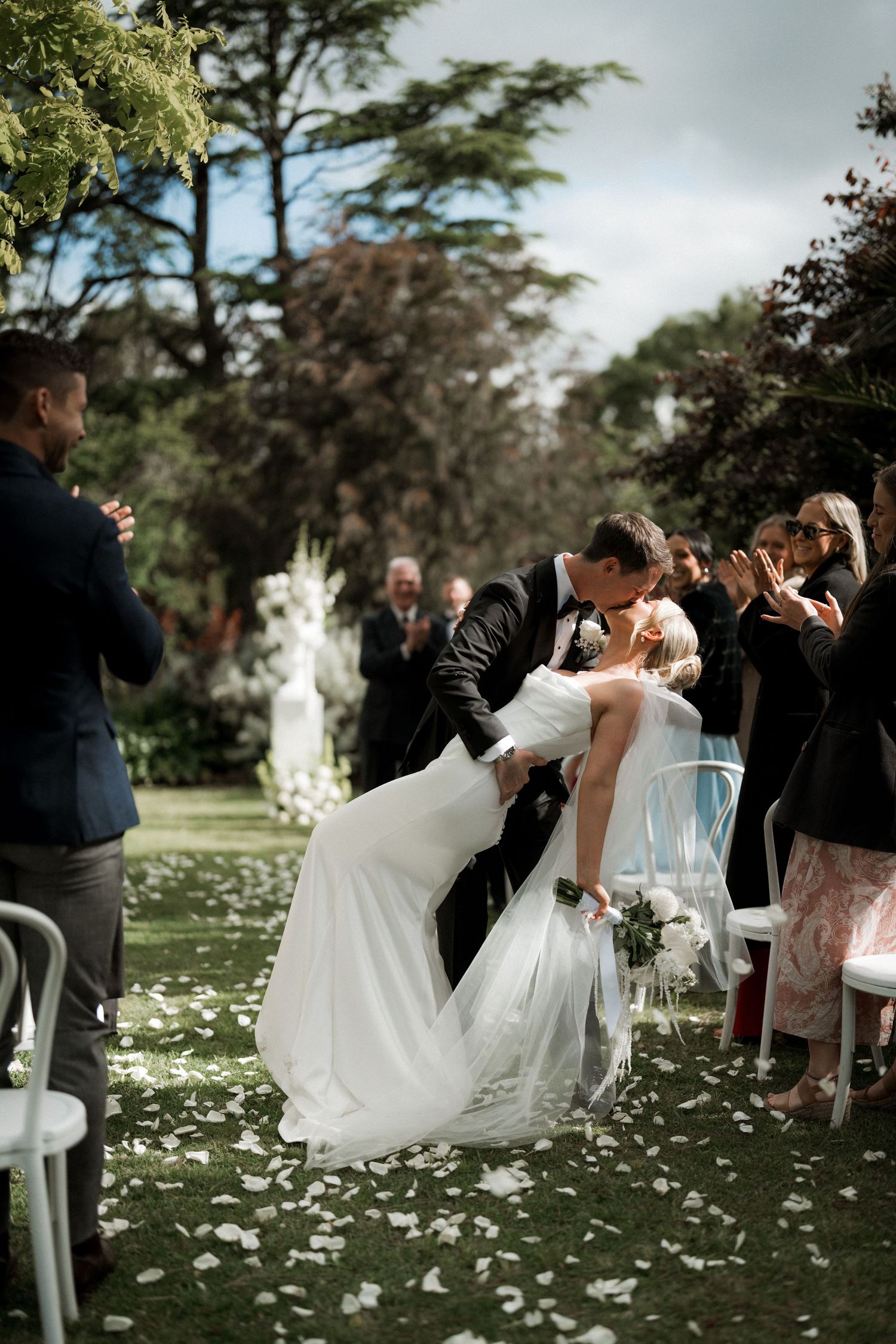 Wedding guests celebrate on a lawn scattered with white flower petals during an outdoor ceremony.