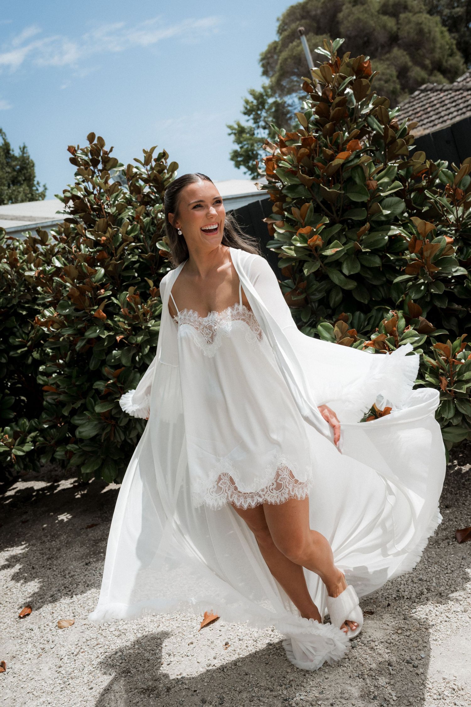 A bride in a white lace dress playfully poses on the ground against a backdrop of magnolia trees.