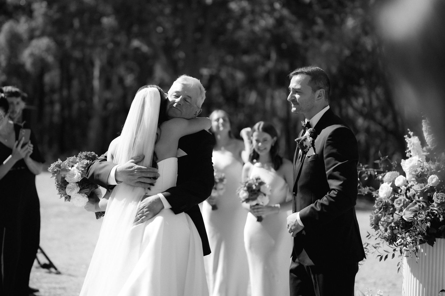 Wedding party members in formal attire pose together with bouquets at an outdoor celebration.