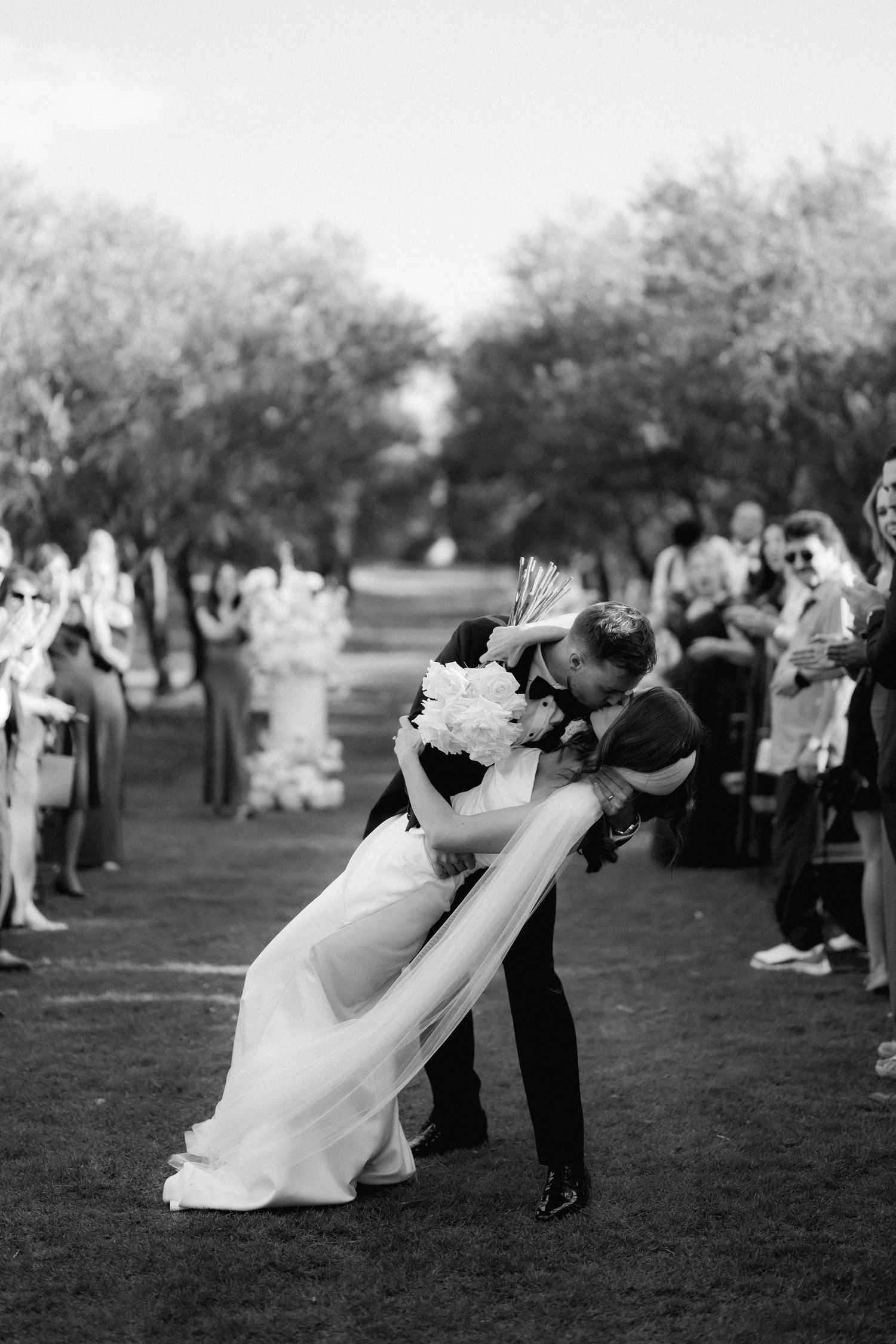 Black and white photo of wedding couples performing synchronized dips on a grassy lawn.