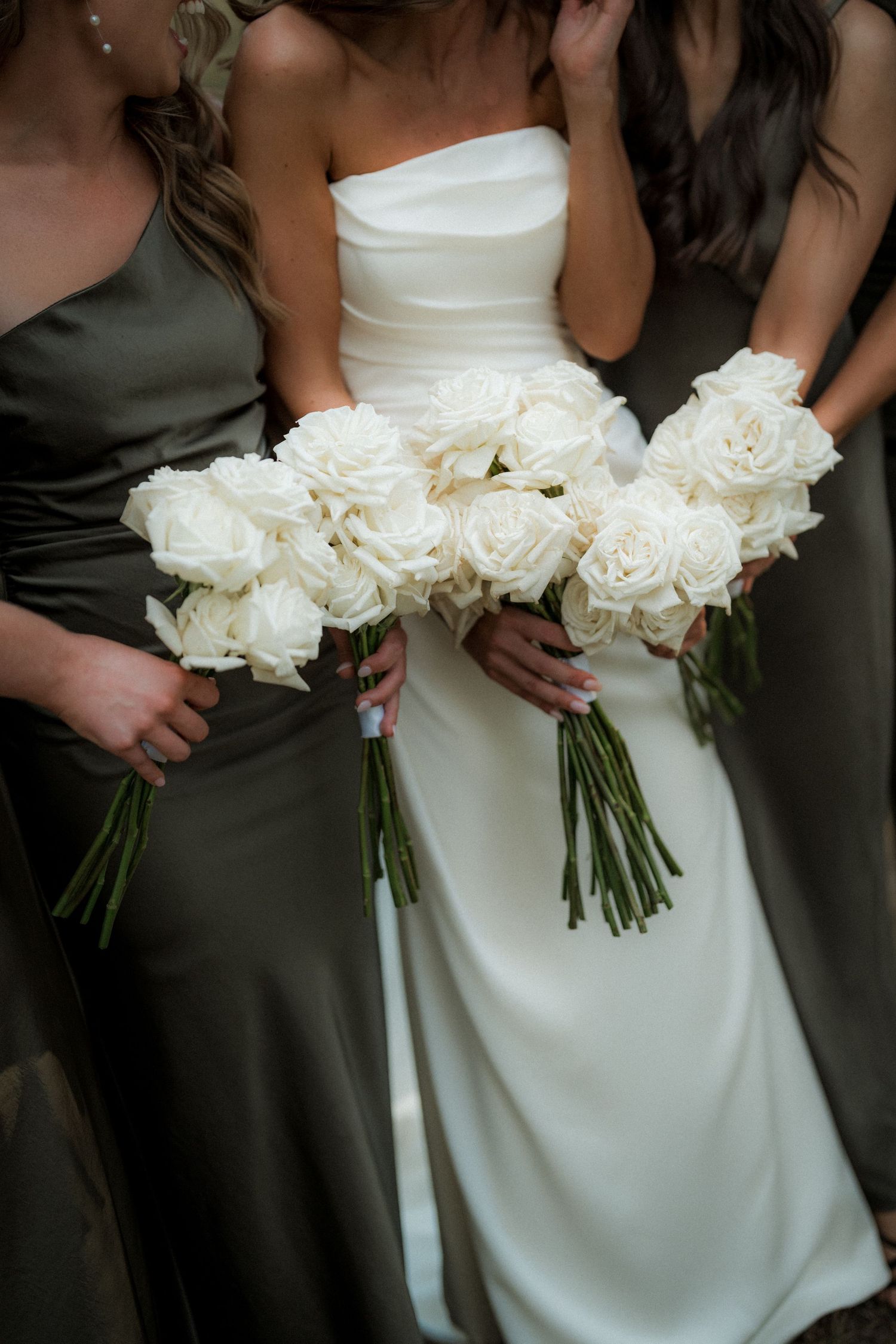 Close-up of white rose bouquets held against white wedding dress and dark bridesmaid dresses.