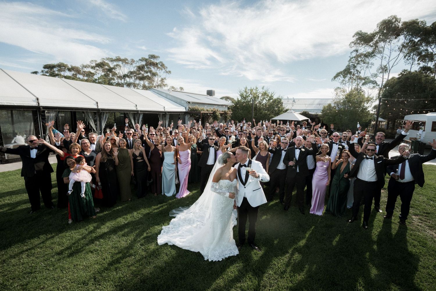 A wedding celebration with guests gathered outdoors on a grassy hillside during a sunny afternoon.