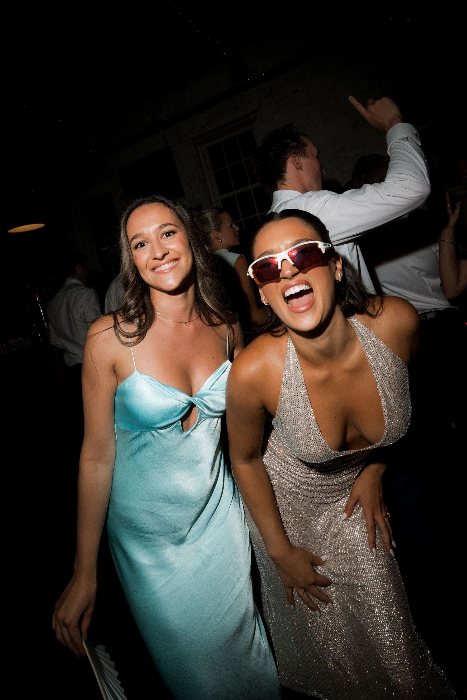 Two women in formal dresses, one in light blue and one in sparkly silver with sunglasses, dancing at a party.