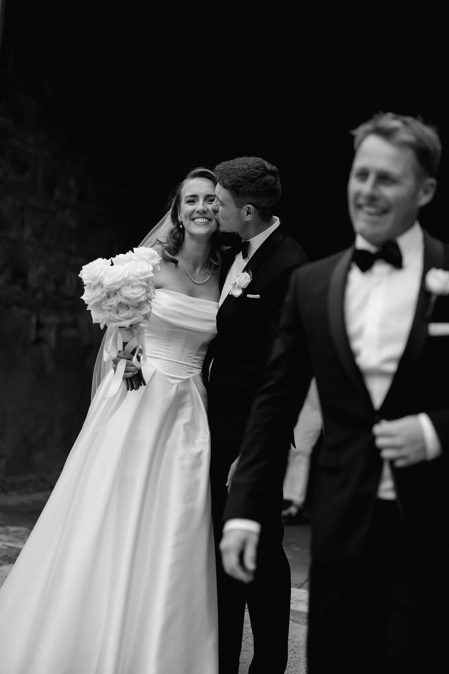 Wedding party members in black tuxedos and white gowns pose together for candid outdoor photos.