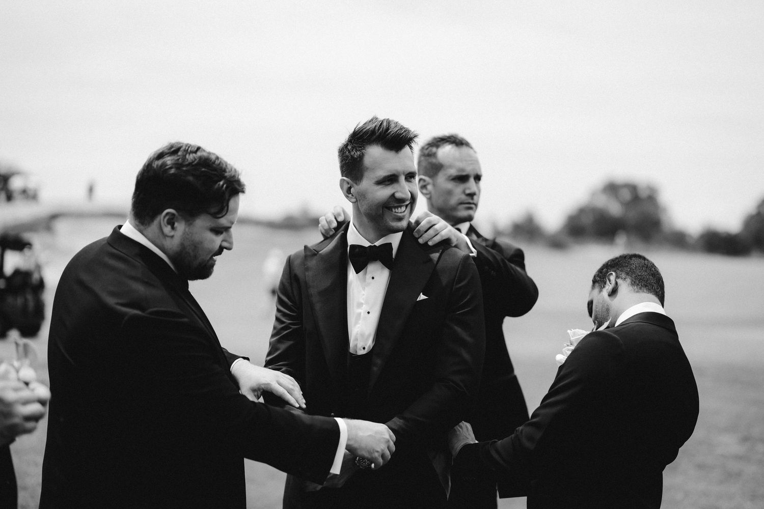 Black and white photograph showing groomsmen helping adjust formal attire before a wedding ceremony.