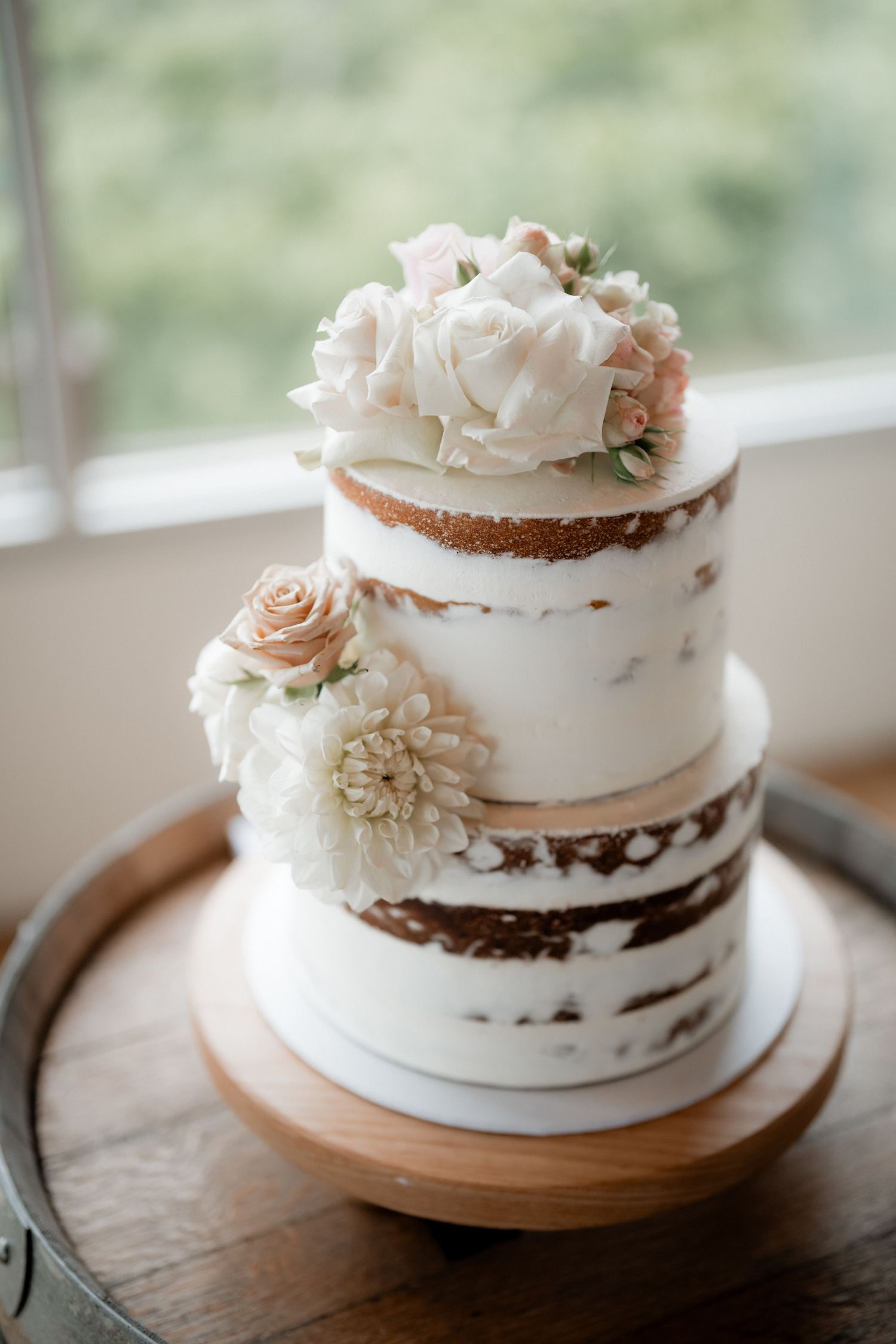 A rustic semi-naked wedding cake with two tiers decorated with fresh white and blush flowers on a wooden cake stand.