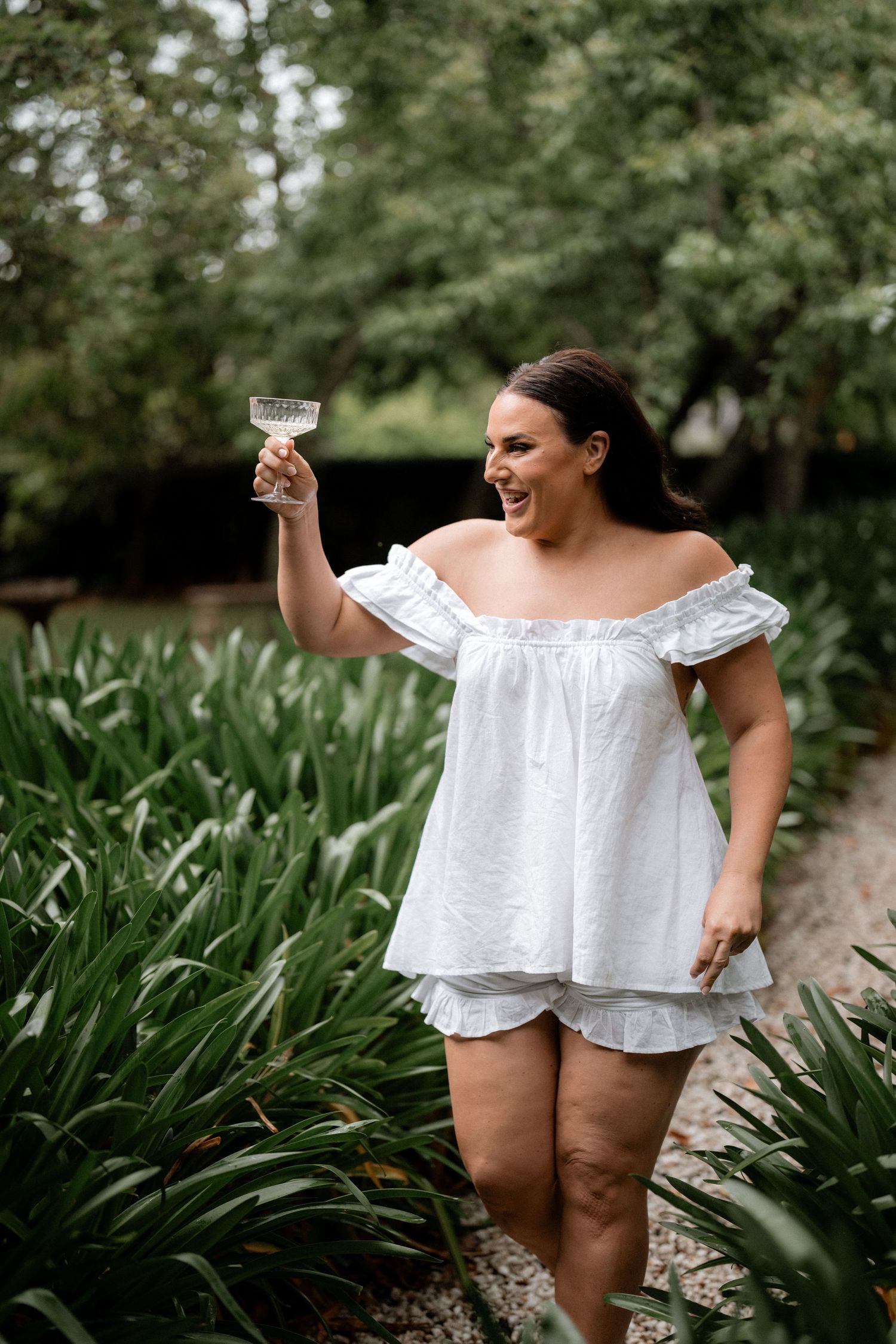Woman in white summer dress raises wine glass while standing in lush garden setting.