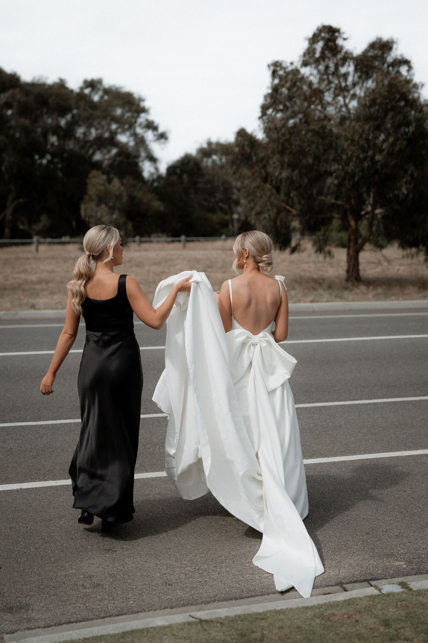 Two women in black and white dresses walking together on an outdoor road near trees.