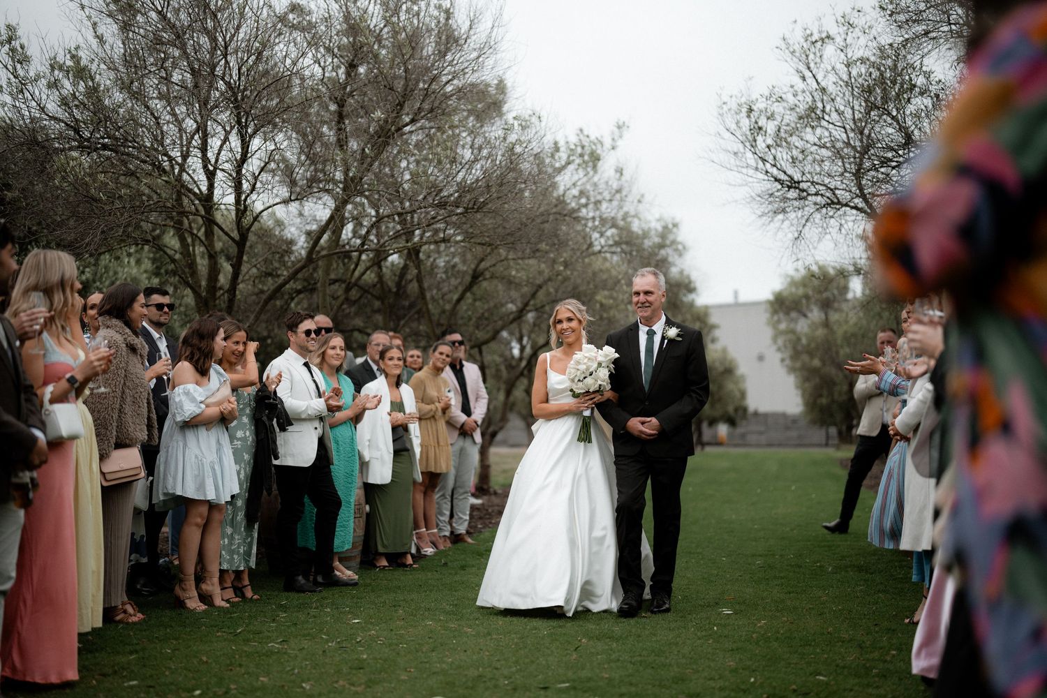 Wedding guests watch as newlyweds walk down a grassy outdoor path under overcast skies.