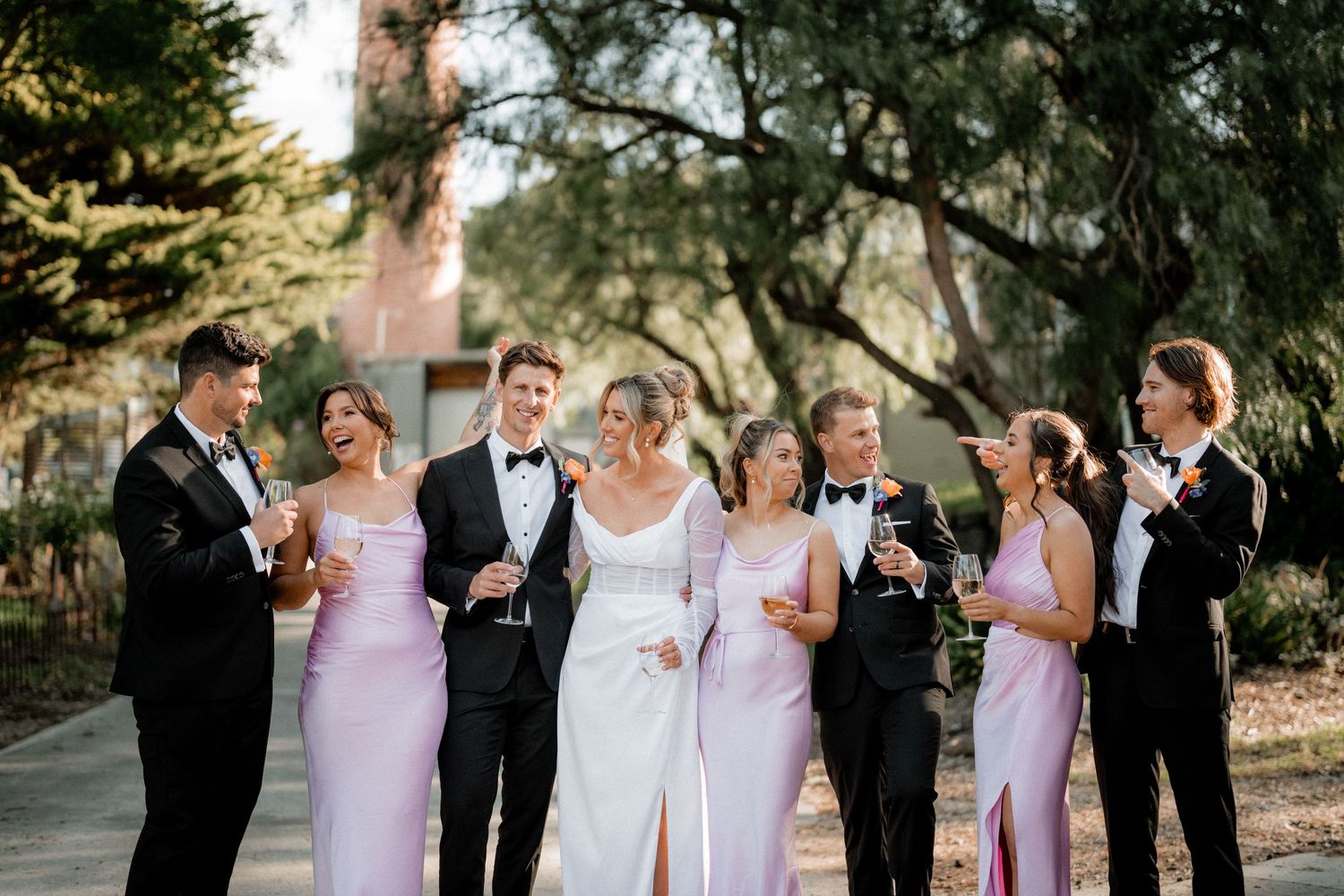 Wedding party in pink dresses and black tuxedos pose together on tree-lined path.