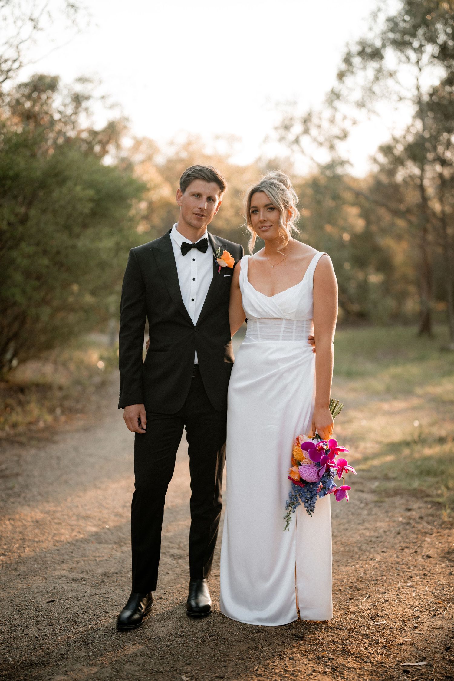 A newlywed couple poses on a rustic path at sunset with the bride holding a vibrant bouquet.