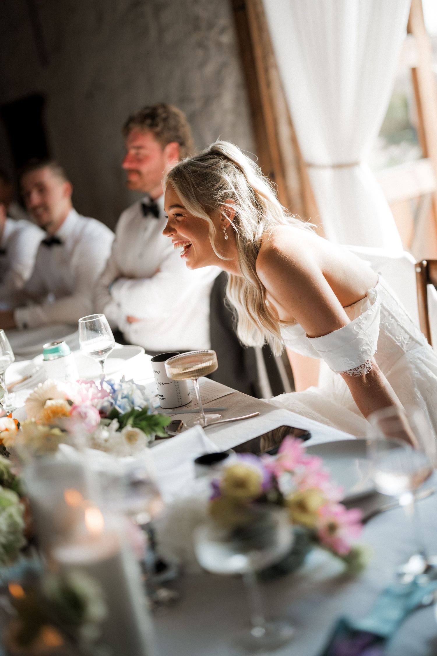 Radiant bride in white gown laughing joyfully at wedding reception head table surrounded by floral centerpieces.