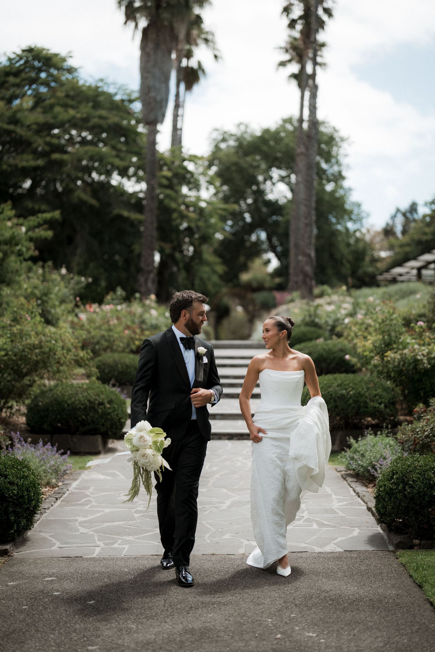Bride and groom walking hand in hand through an elegant garden pathway lined with palm trees and hedges.