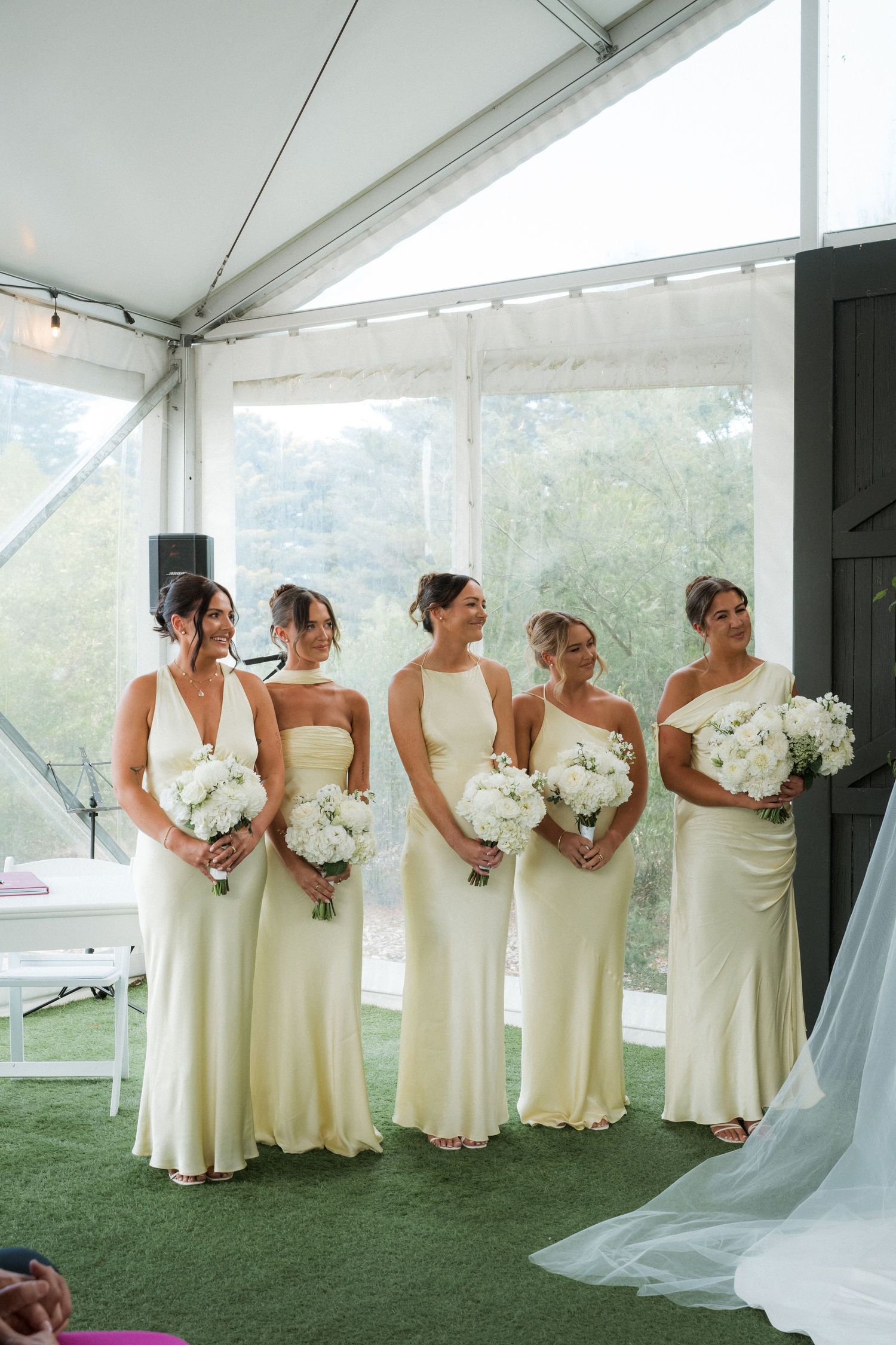 Bridesmaids in cream-colored dresses line up holding white bouquets under a white tent.
