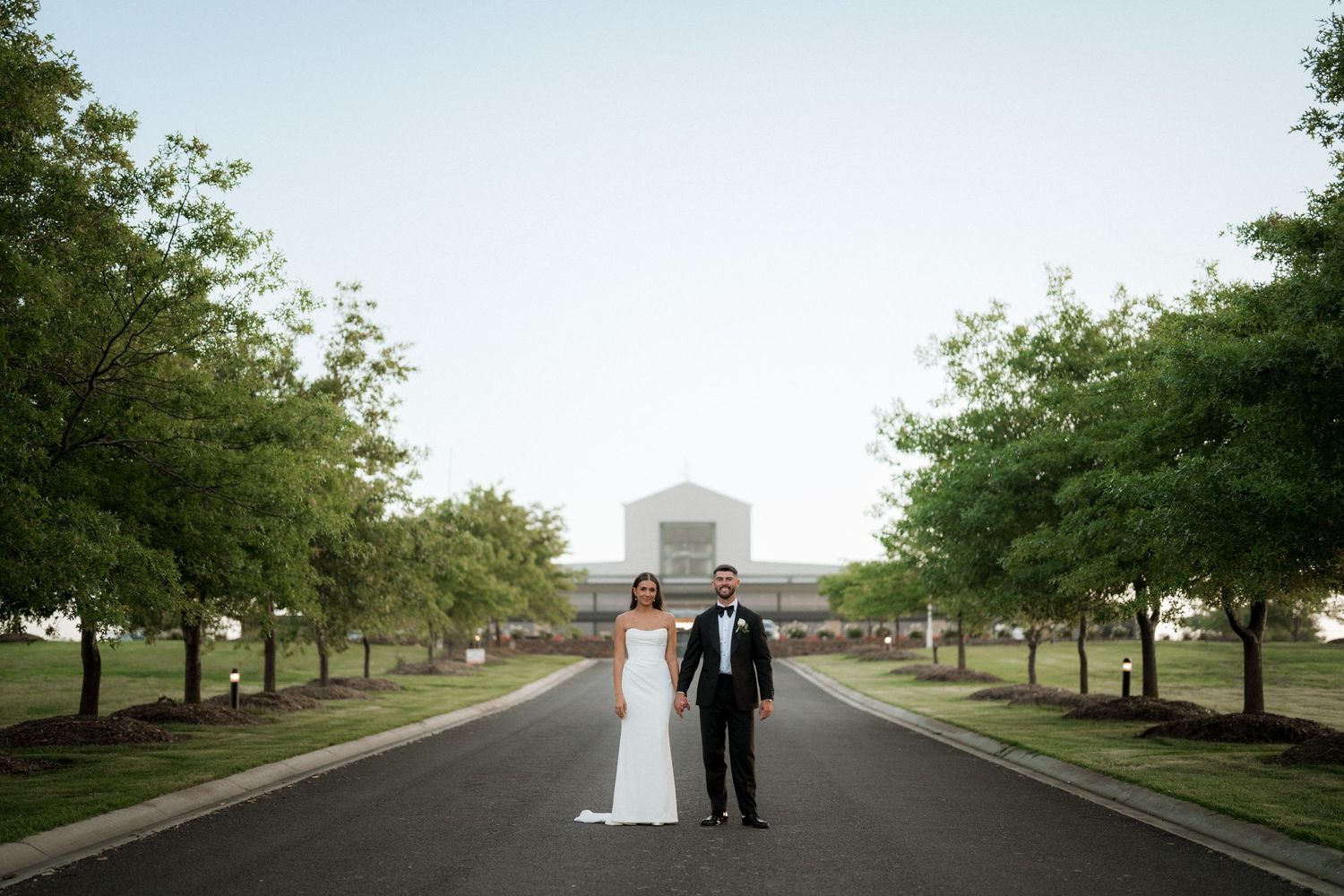 A couple stands on a tree-lined pathway with a basketball hoop visible in the distance.