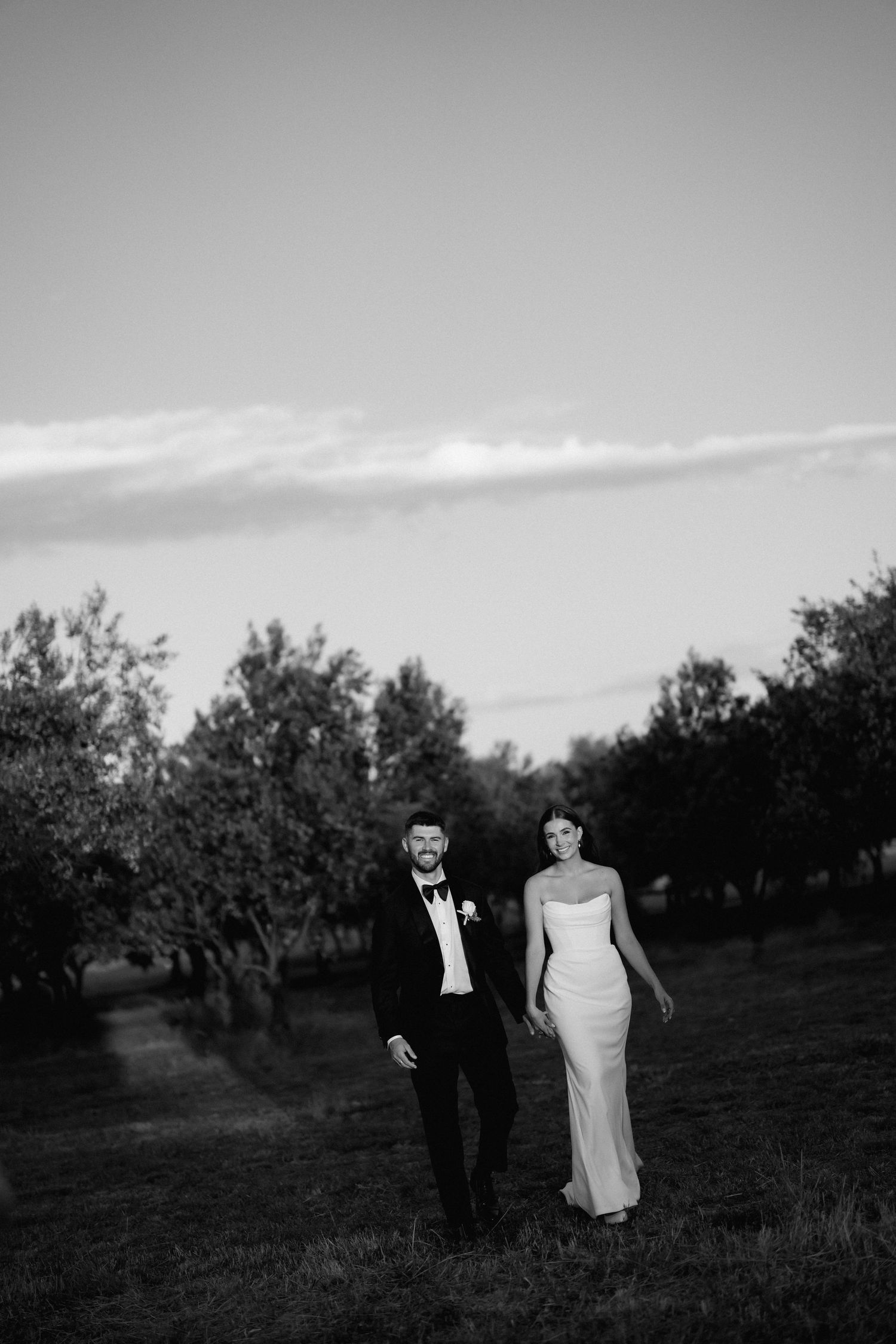 Black and white photo of a couple walking through an orchard in formal attire.