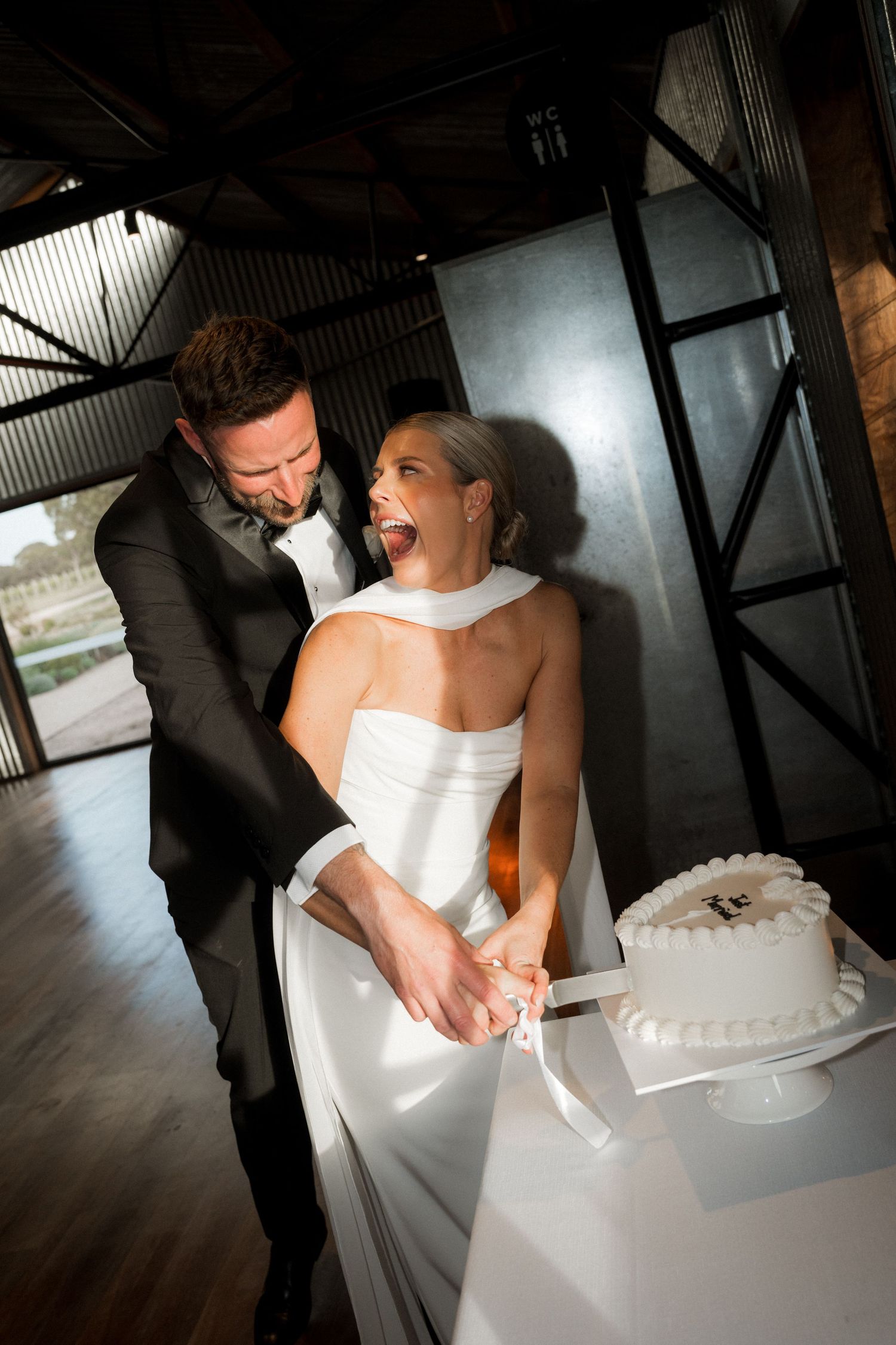 Couple in formal attire cutting their white wedding cake together at reception venue.