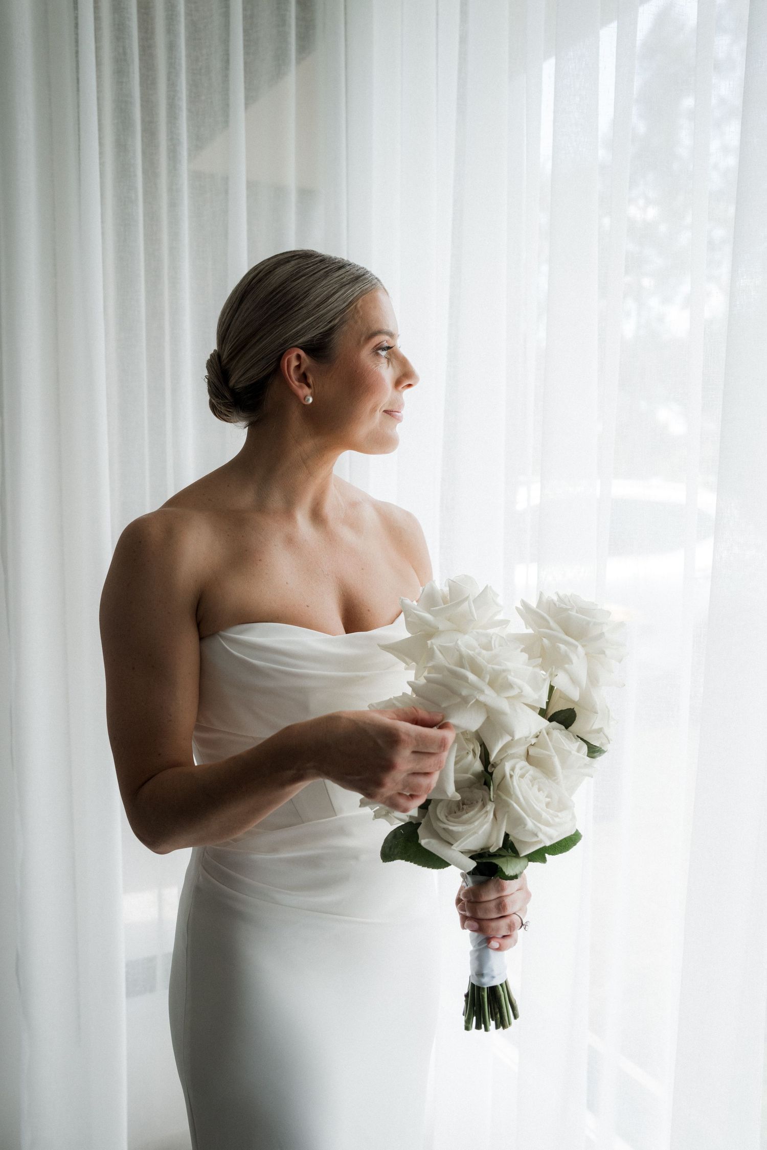 Elegant white wedding dress and bouquet against sheer curtains in natural light.
