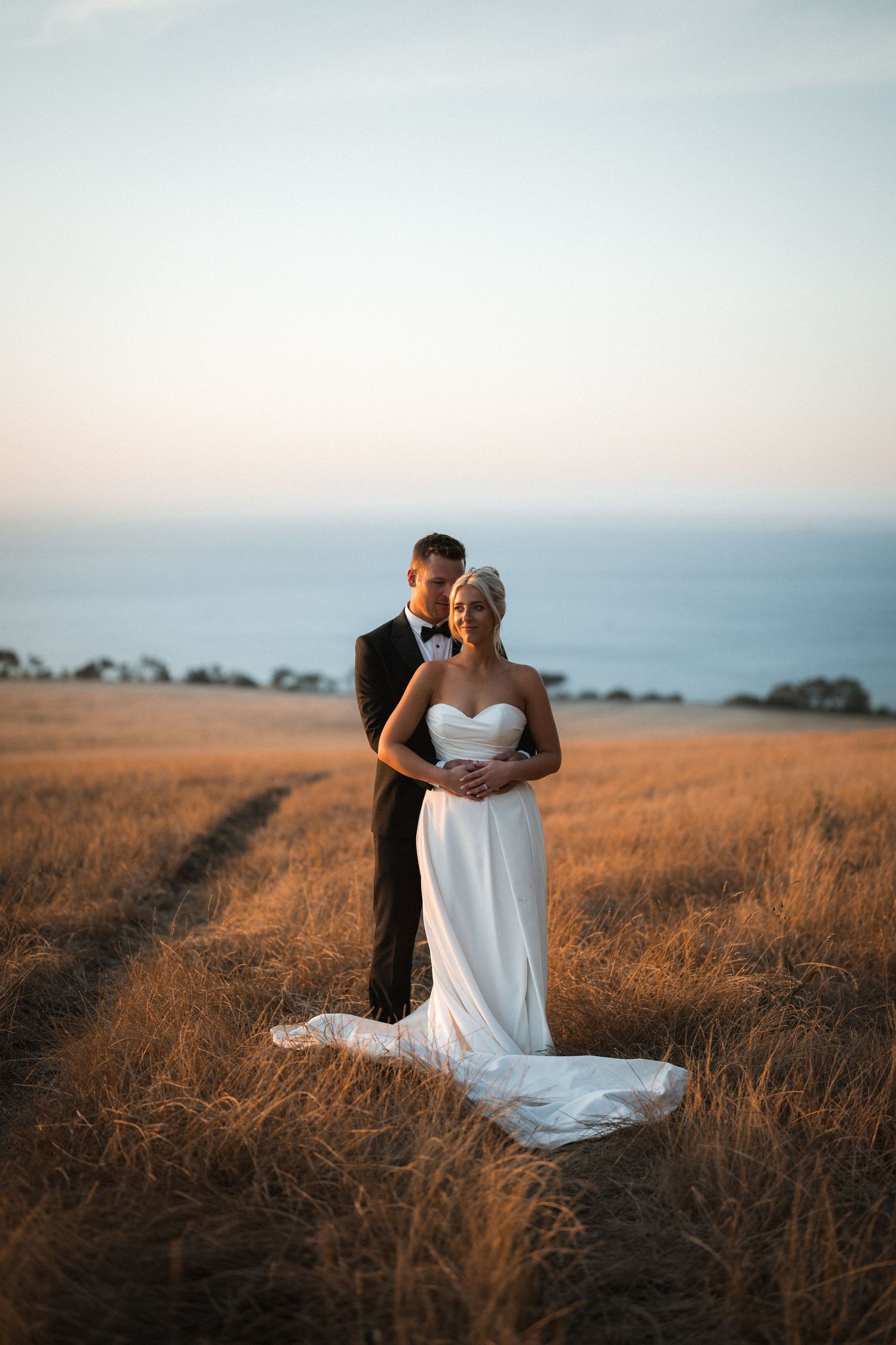 Newlywed couple embraces in golden field at sunset with ocean view in background.