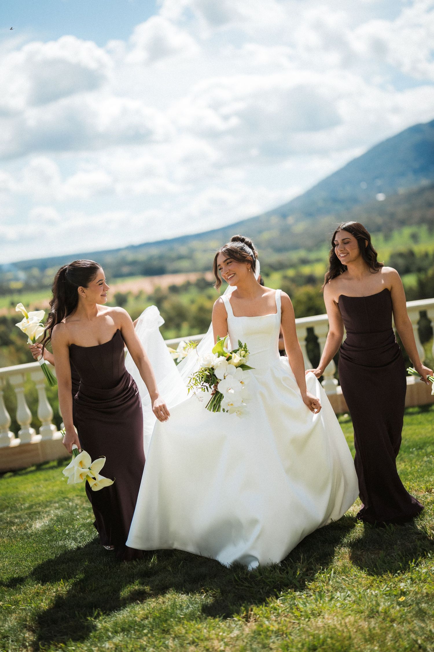 Bride in a white wedding gown walks on a grassy hillside with two bridesmaids in dark dresses holding white lily bouquets.