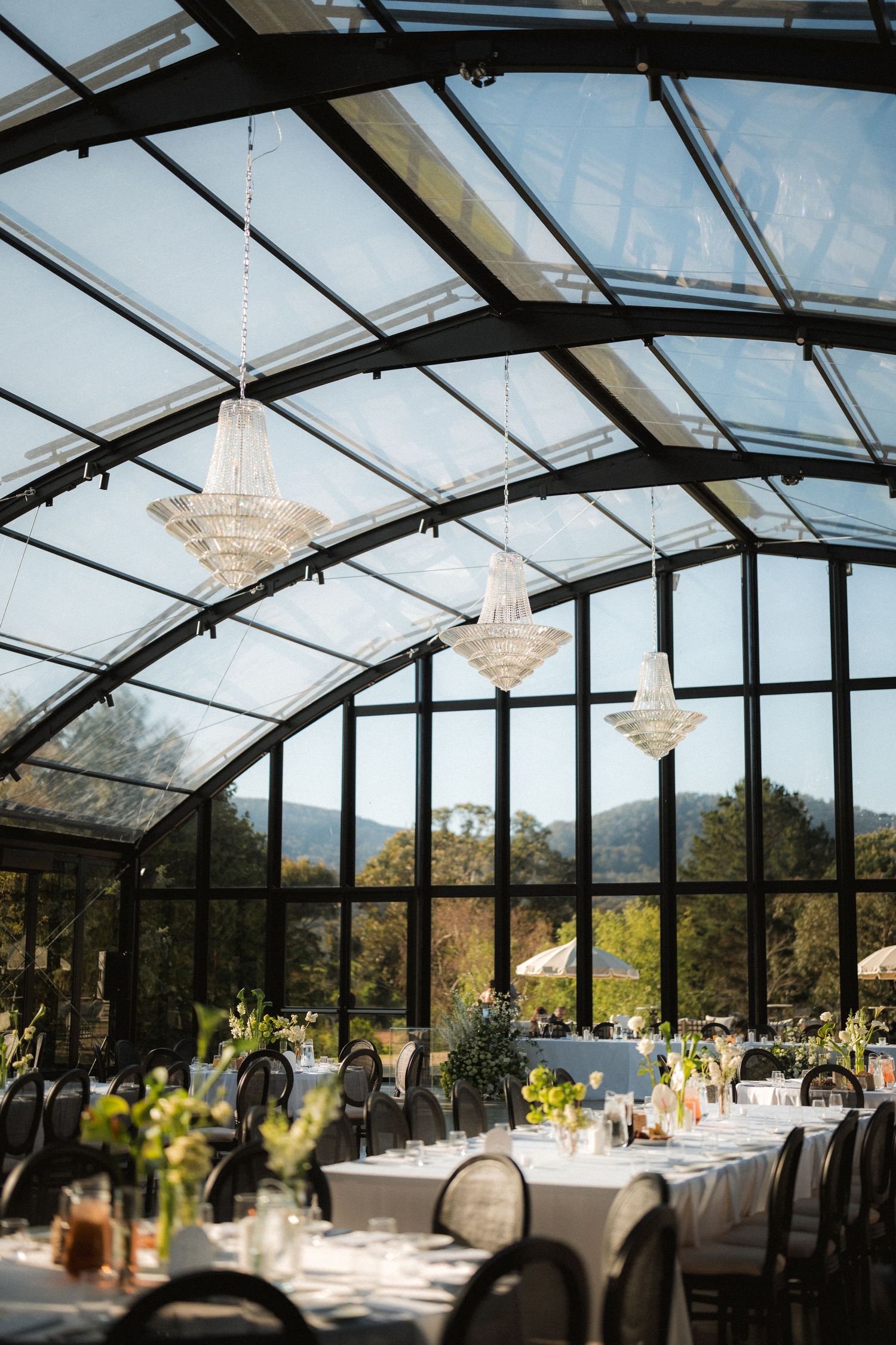 Elegant glass-walled wedding venue with crystal chandeliers, set banquet tables, and floral centerpieces overlooking scenic mountain views.