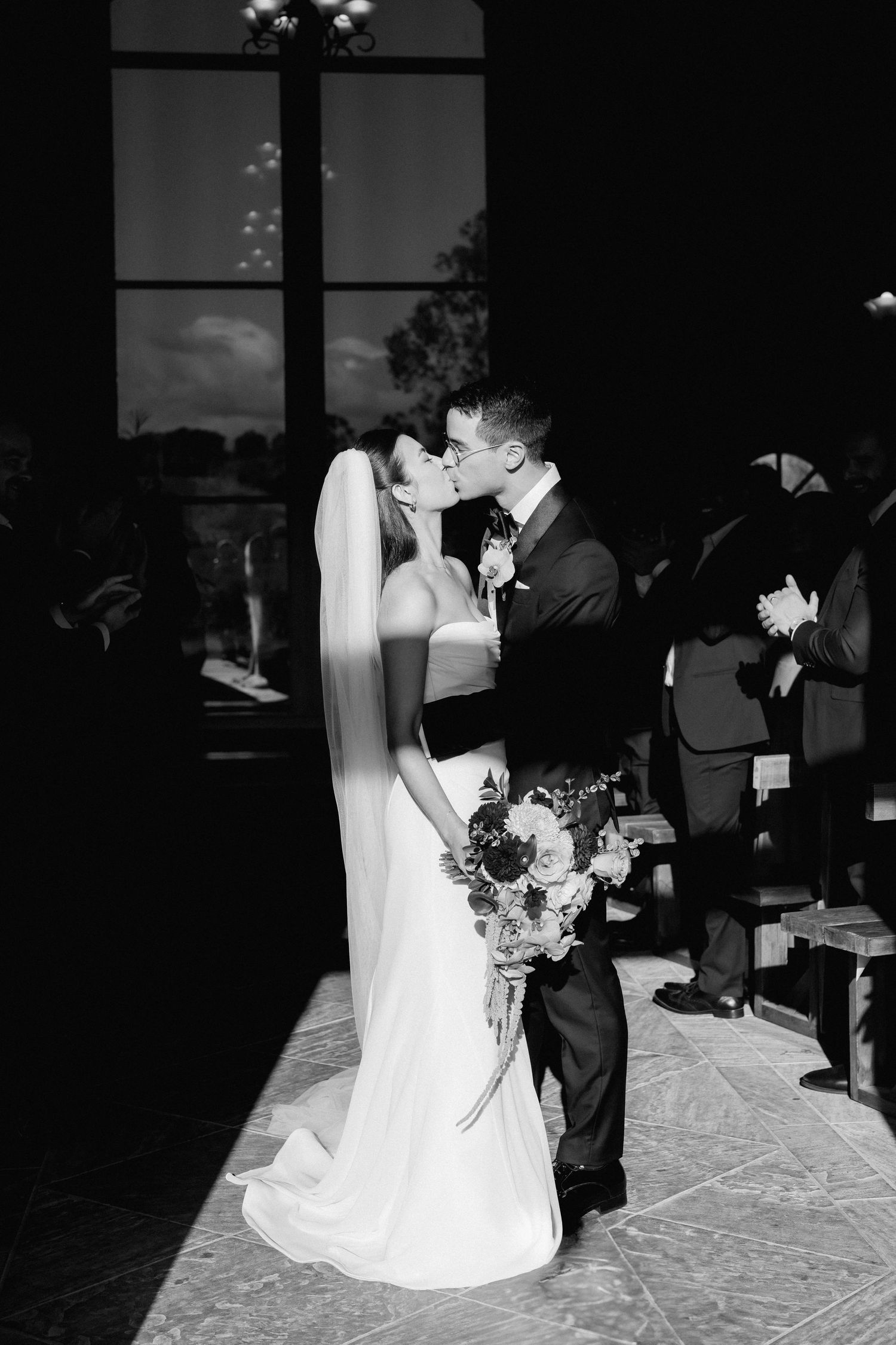 Black and white photo of bride and groom sharing first kiss at wedding ceremony altar.