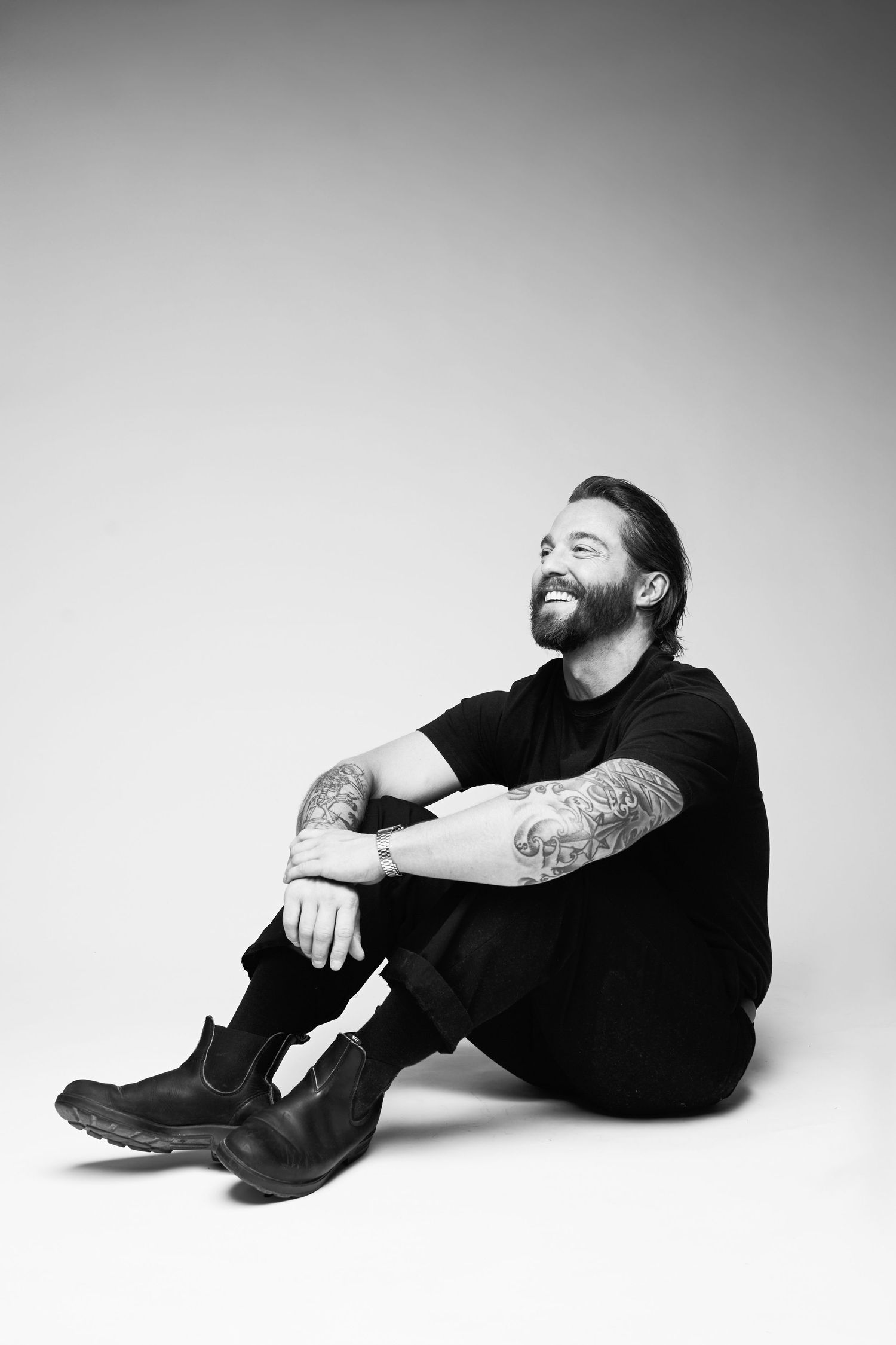 Black and white photo of a smiling bearded tattooed man sitting on floor in black outfit and boots.