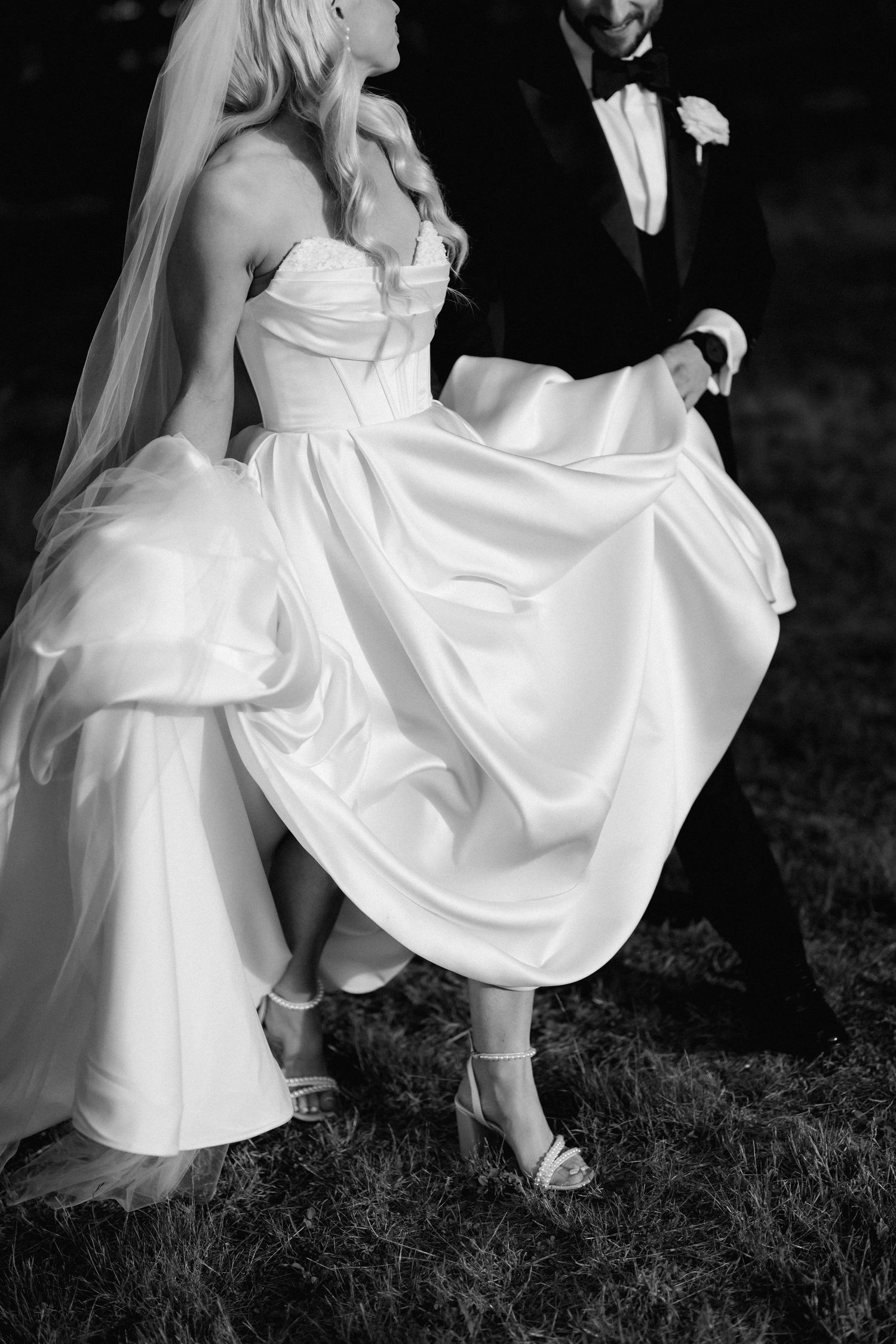Black and white photo of a bride in a flowing satin gown and veil, lifting her dress while walking with a groom in a tuxedo.