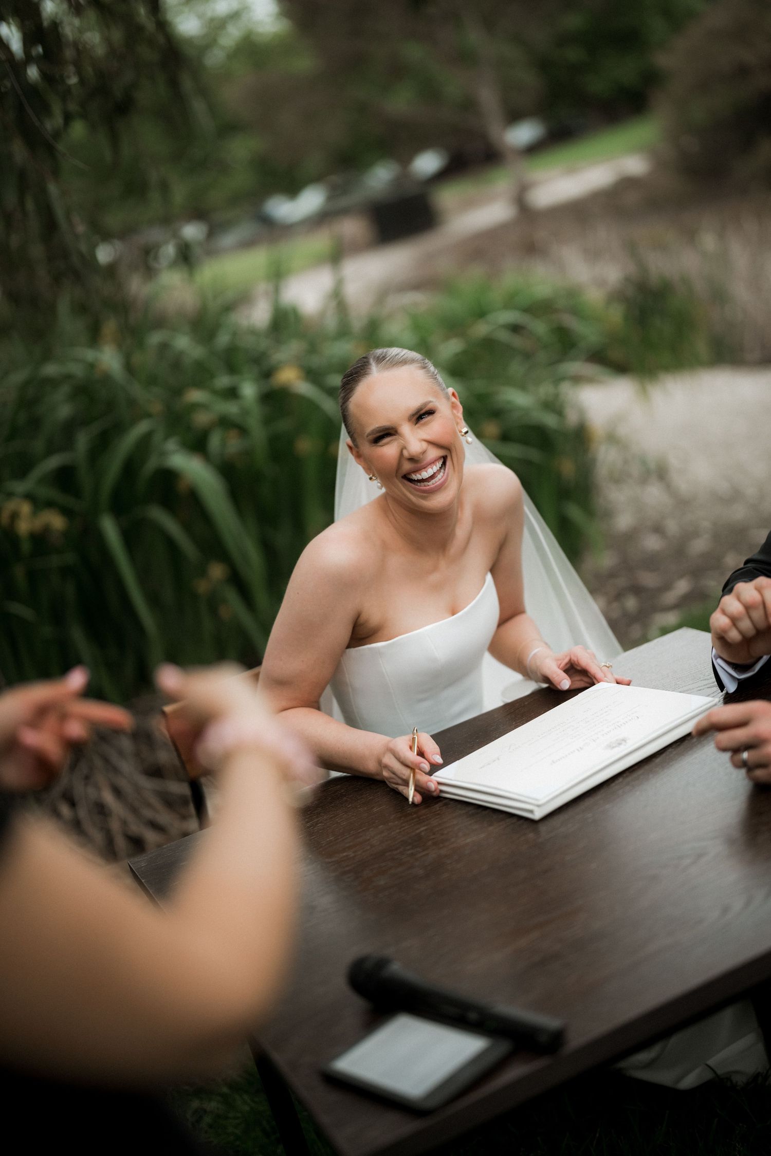 Smiling bride in strapless gown and veil signs marriage register at outdoor wooden table.