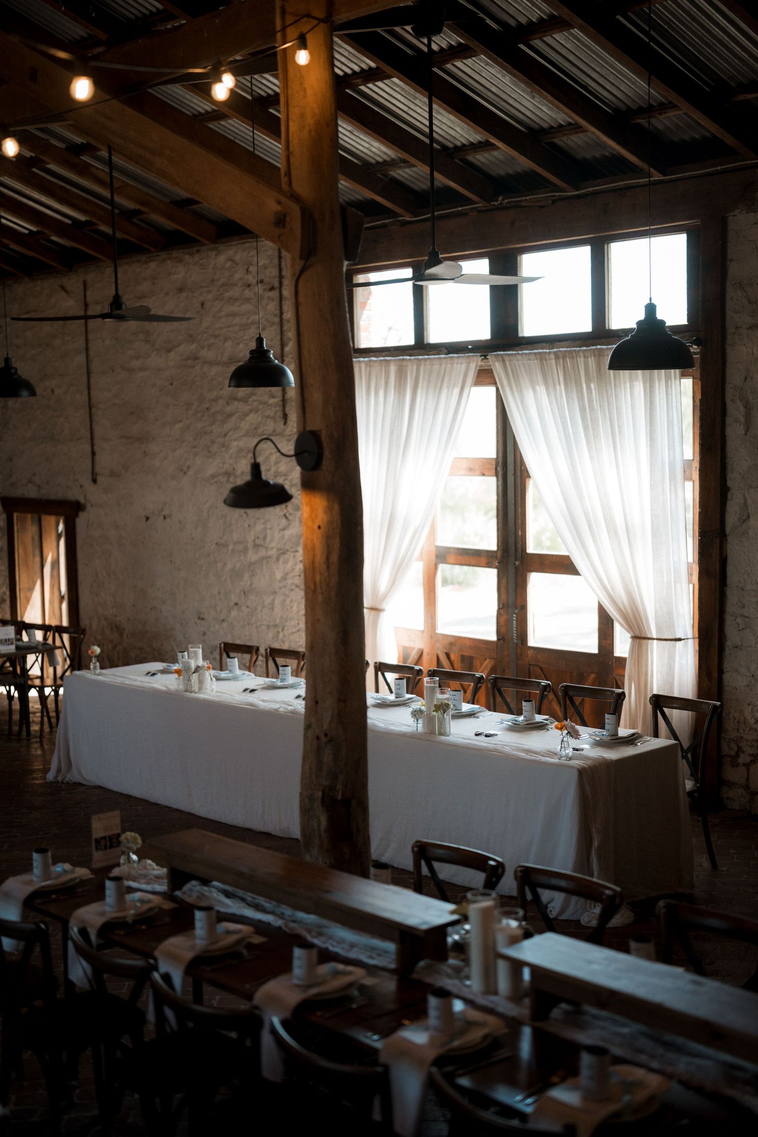 Rustic wedding reception venue with white tablecloths, Edison bulb lighting, and sheer curtains on windows.