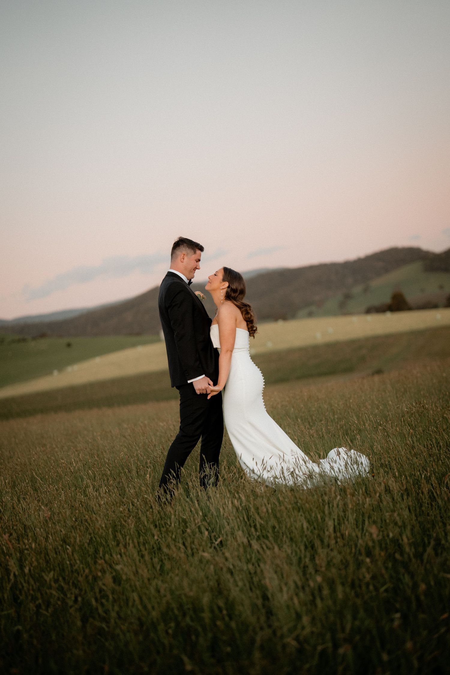 A couple shares a romantic kiss in a serene field at dusk with rolling hills in the background.