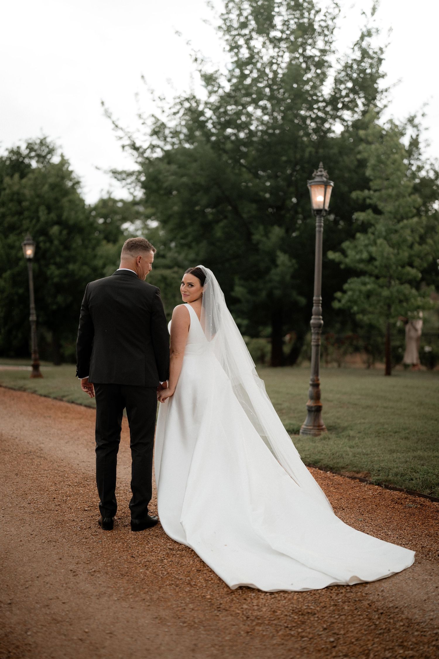 Wedding couple walks along a tree-lined path with the bride's elegant white dress trailing behind.