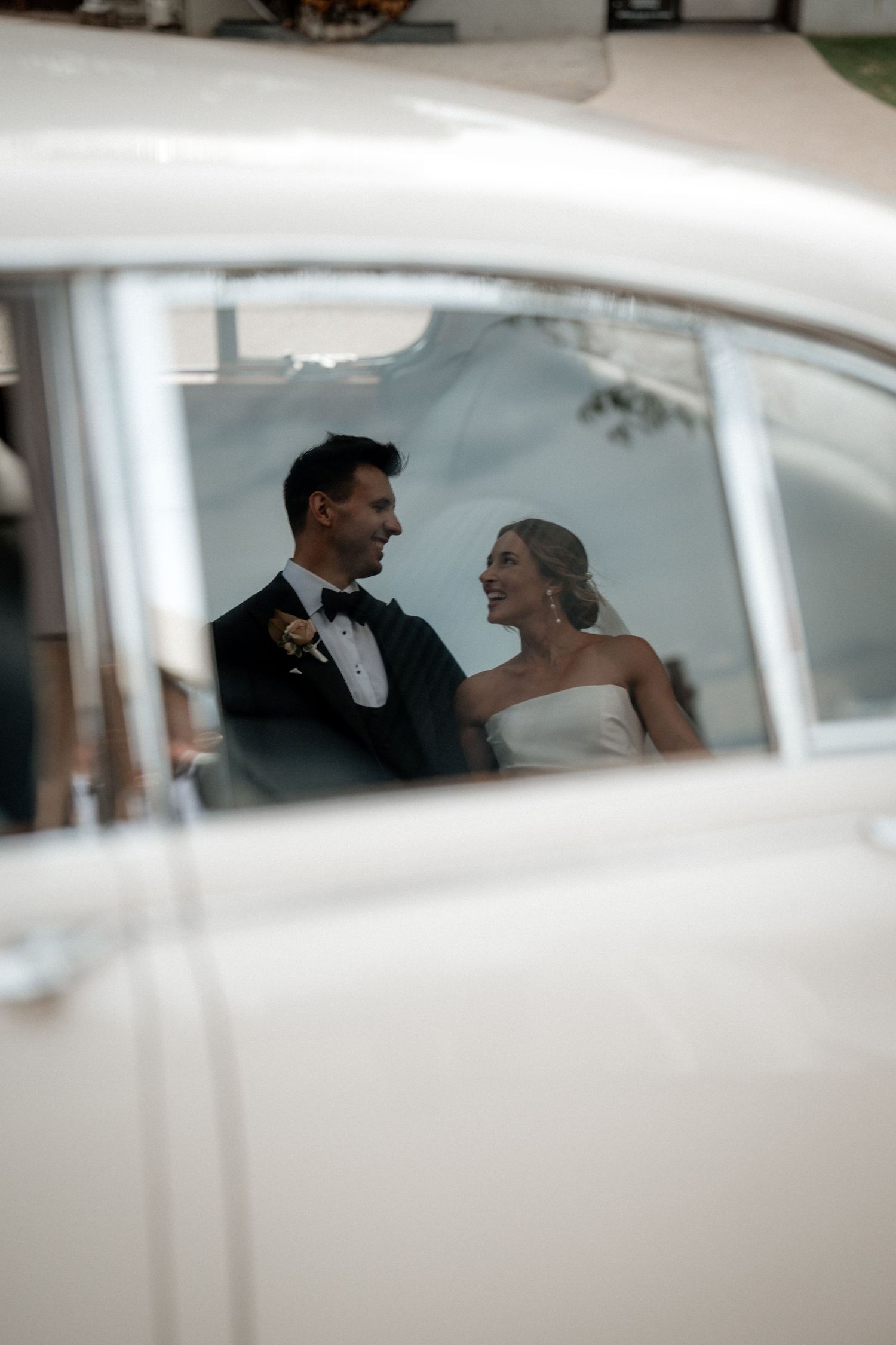 A bride and groom share a joyful moment reflected in a classic car's window on their wedding day.