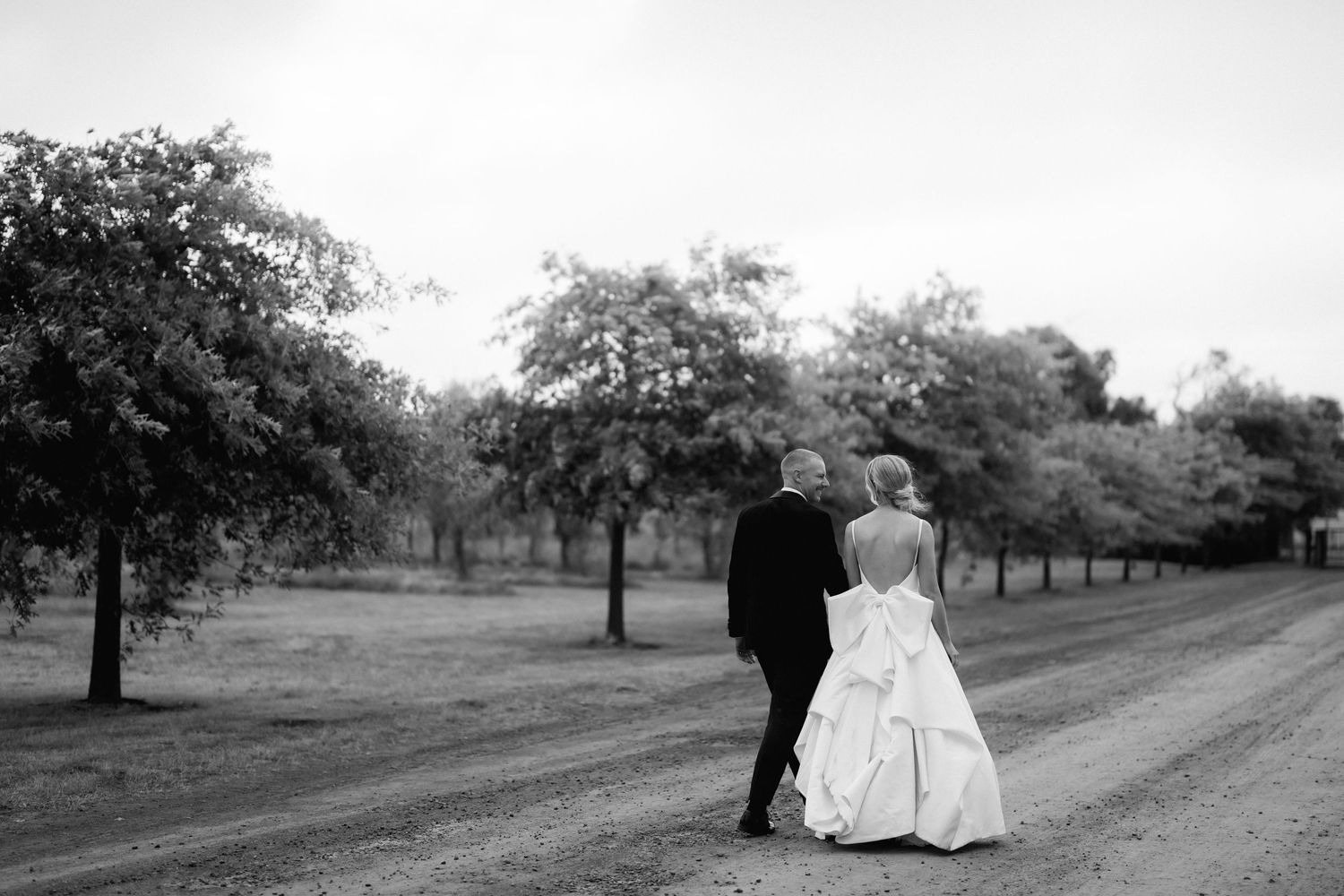 A couple in wedding attire walks along a tree-lined path in black and white.