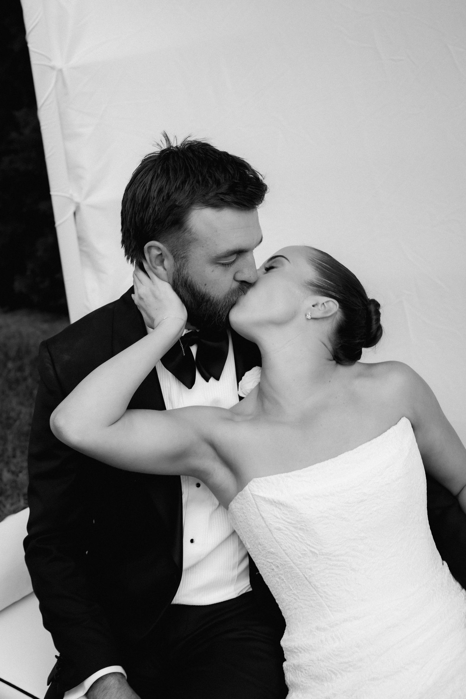 Black and white close-up of bride and groom sharing a passionate kiss while seated at their wedding reception.