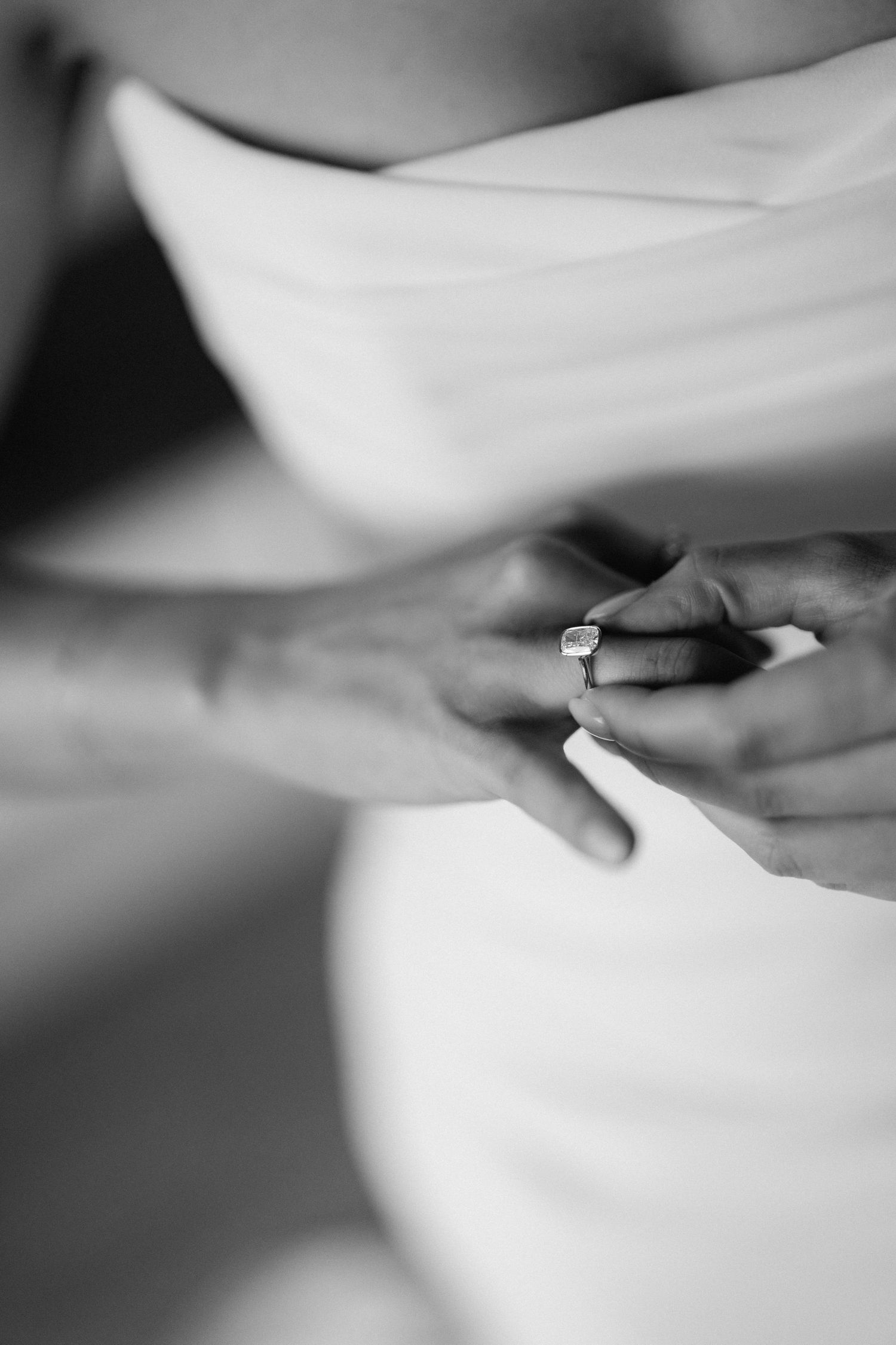 Close-up black and white photo of bride wearing elegant square diamond engagement ring in white dress.