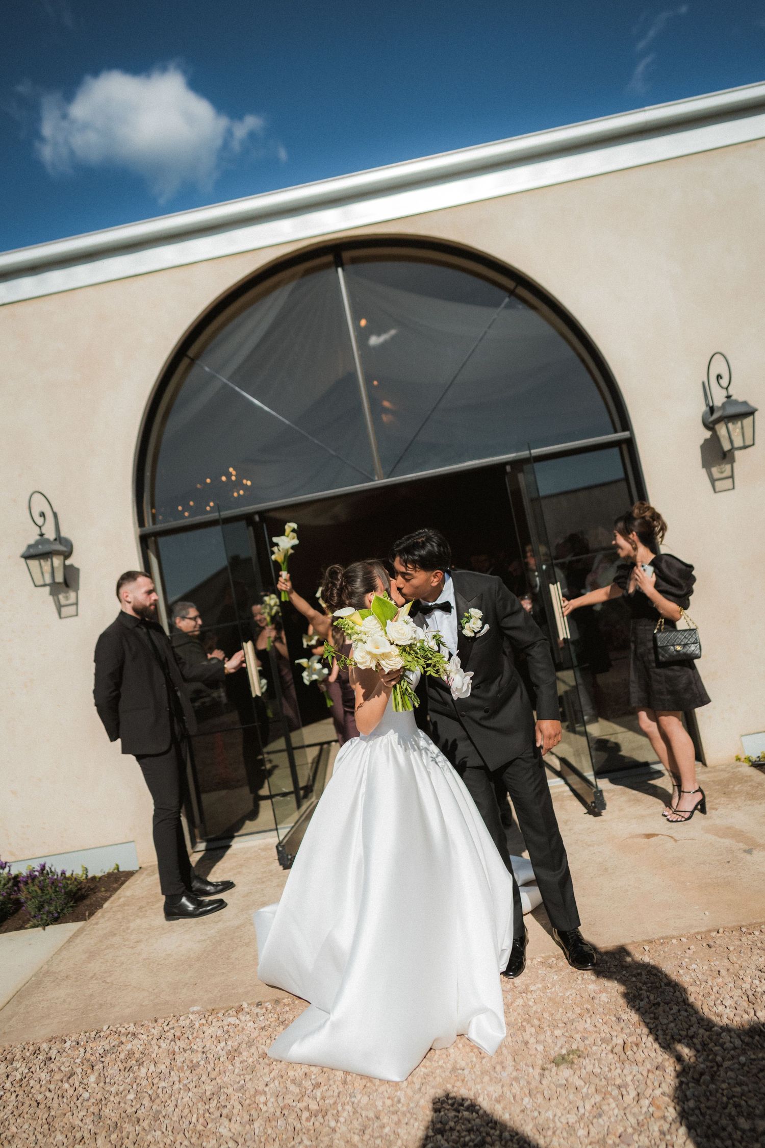 Bride and groom kiss outside an arched glass doorway while guests in formal attire cheer and toss petals on a sunny wedding day.